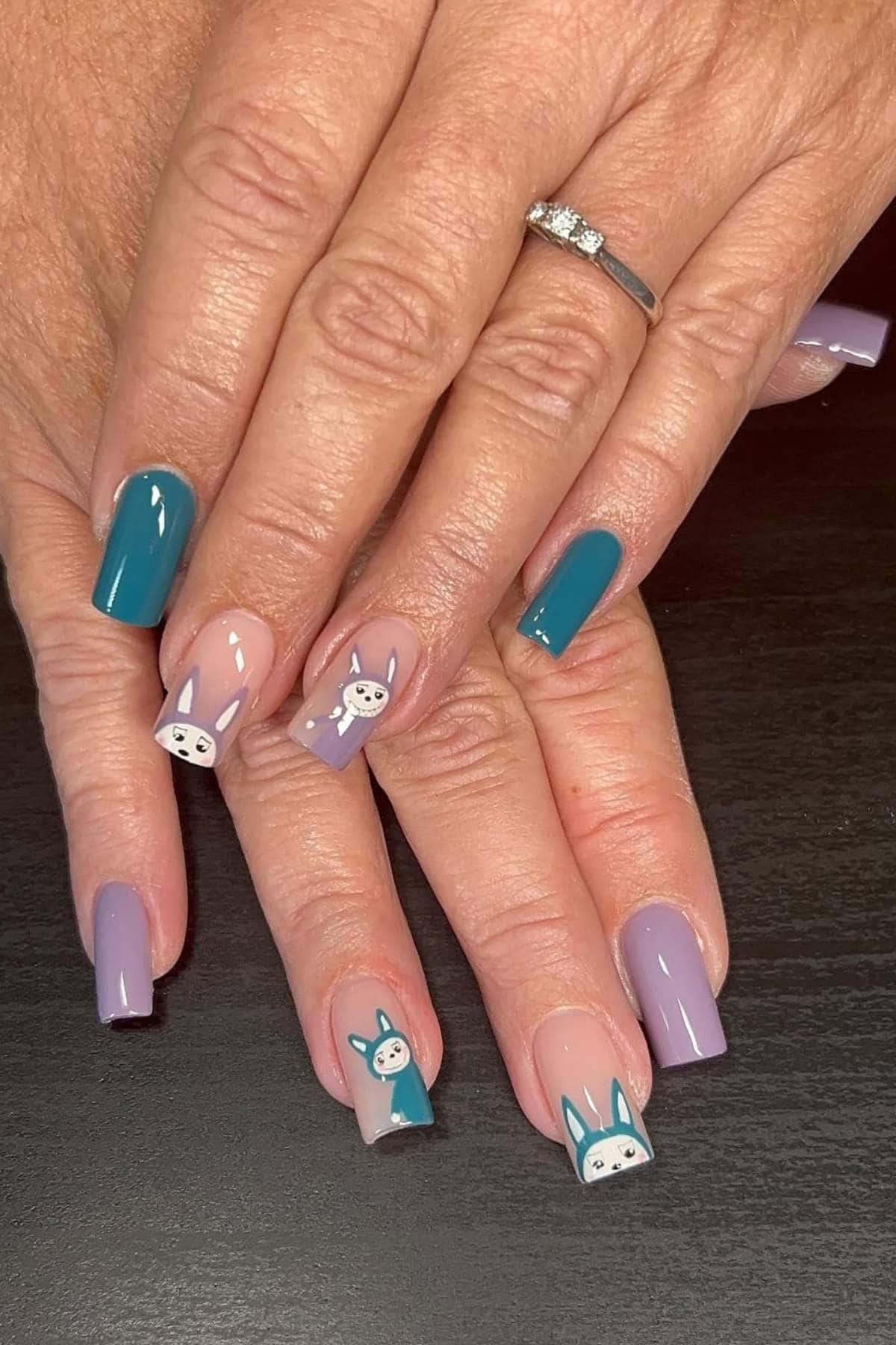 A hand with teal and lavender manicured nails, some featuring cartoon animal faces, resting on a dark surface.