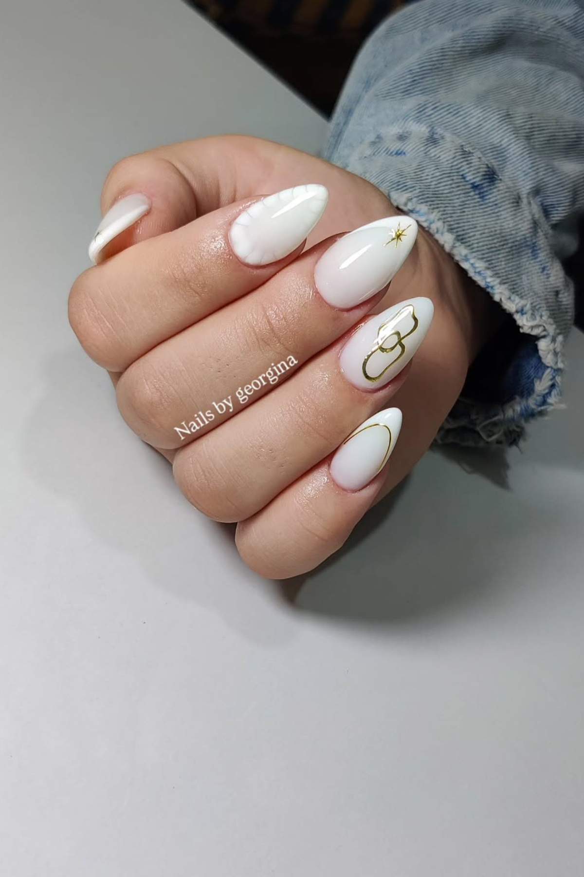 A hand with almond-shaped white nails featuring subtle gold designs and a denim sleeve visible in the background.