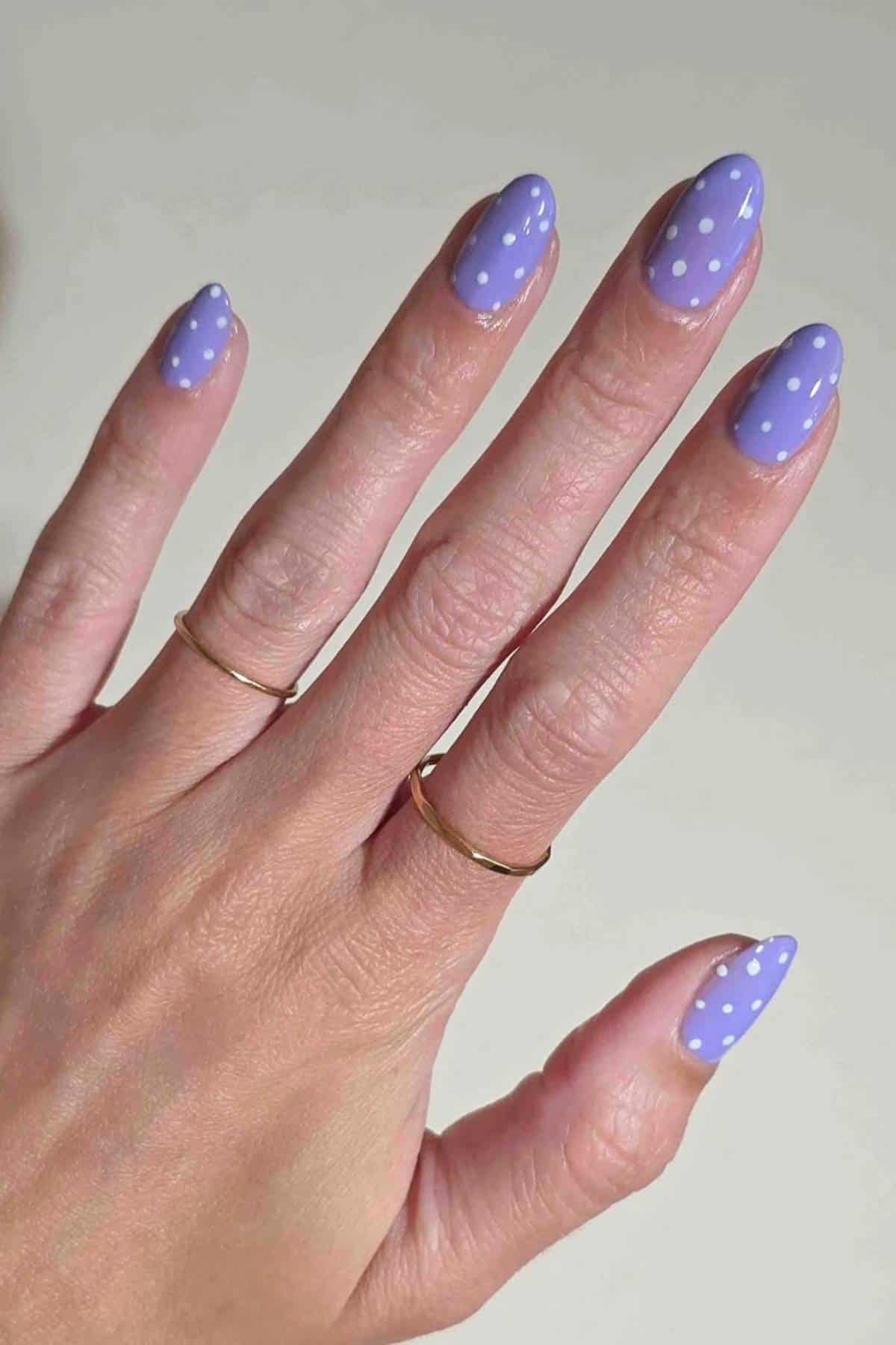 A hand with light purple nail polish and white polka dots on the nails, wearing two thin gold rings on the middle and ring fingers.