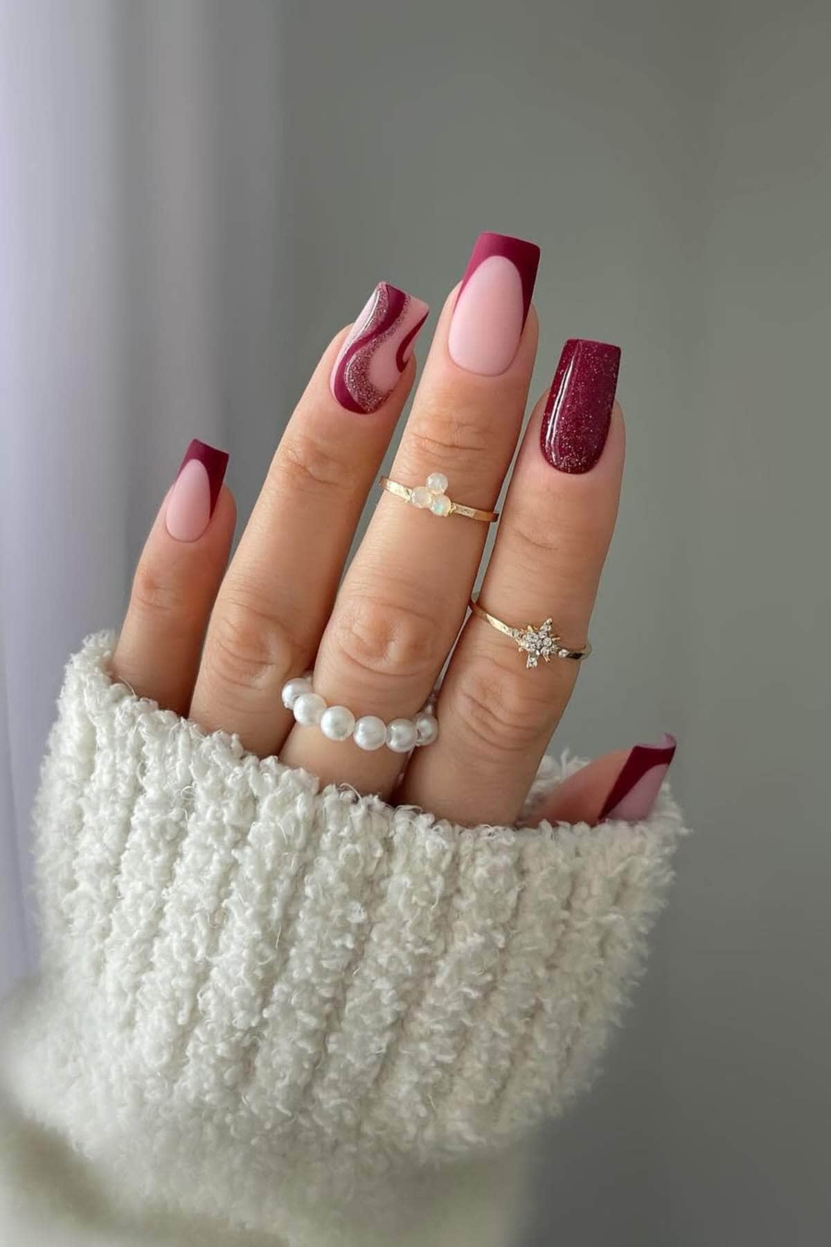 A hand with long, square-shaped nails features a burgundy and pink manicure with various designs, and is adorned with gold and pearl rings. The person wears a white knit sweater.