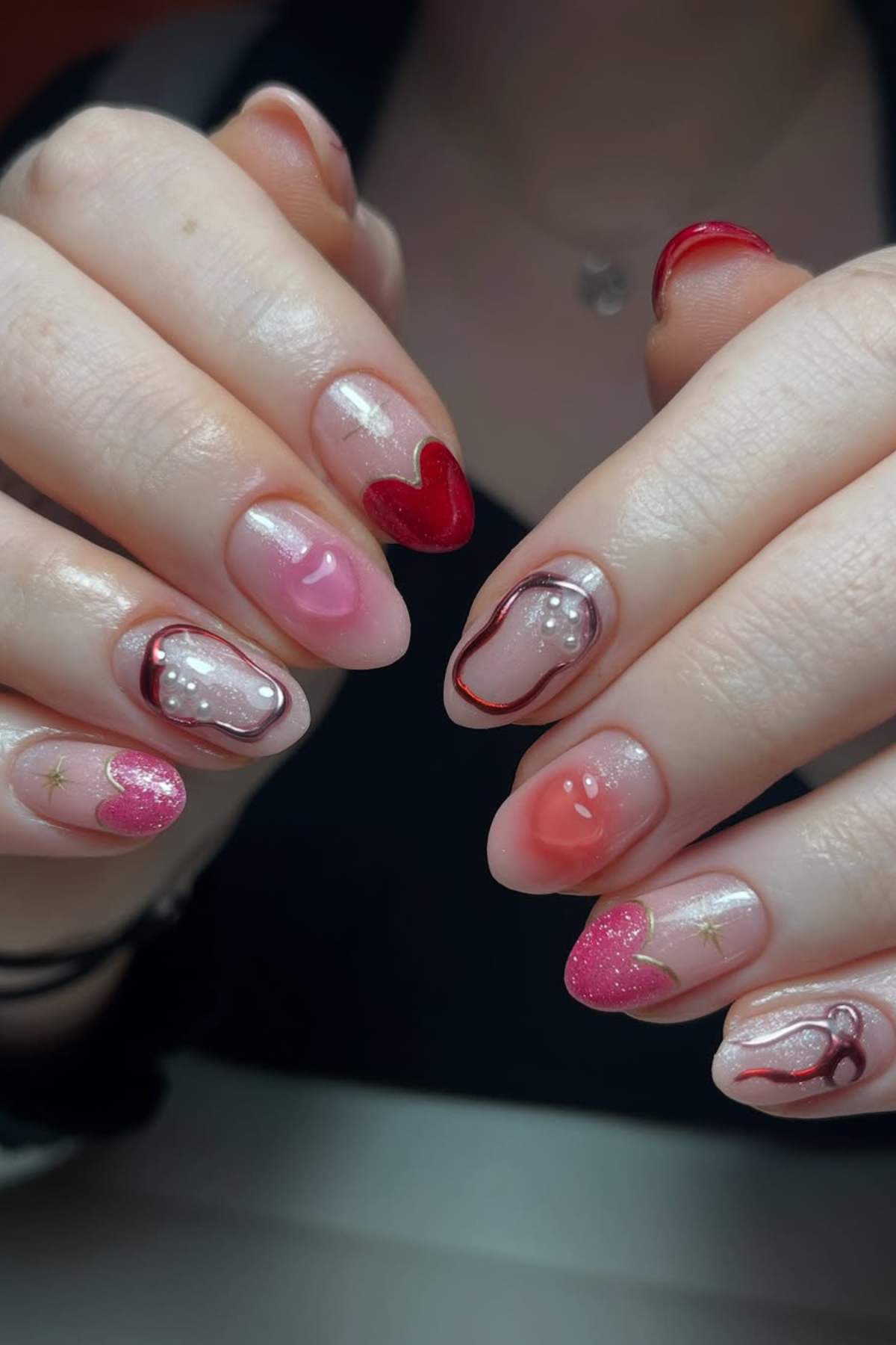 Close-up of hands with almond-shaped nails featuring a Valentine's Day-themed manicure, including red and pink hearts, swirls, glitter, and small pearl accents.