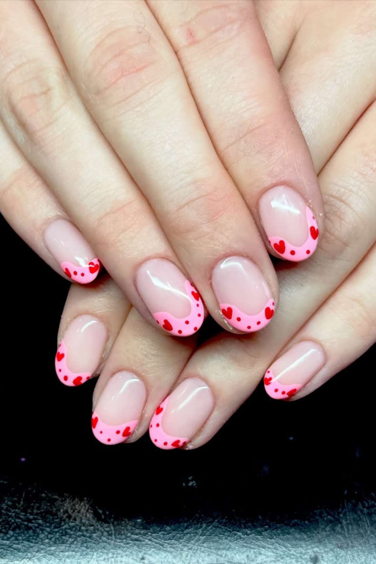 Hands with a pink French manicure featuring red heart and dot designs on the tips of each fingernail.