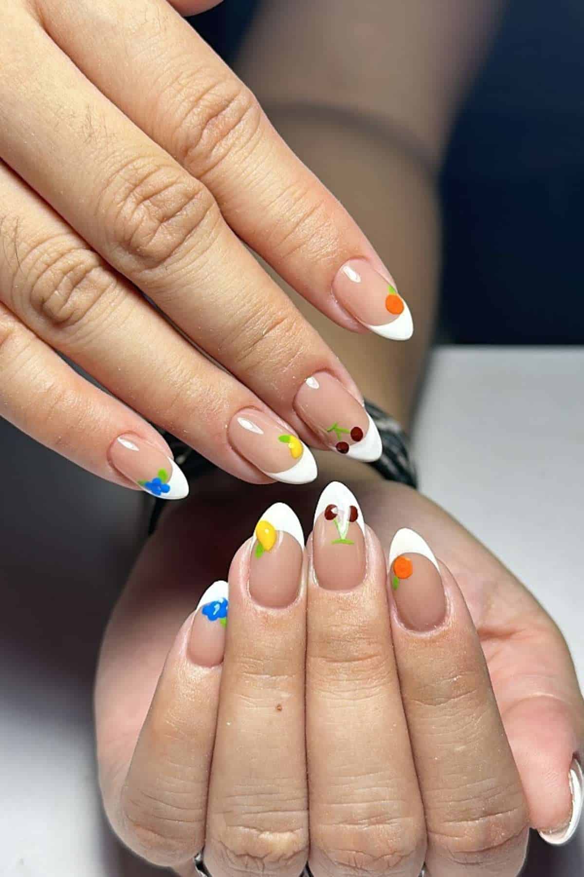 Hands with almond-shaped nails featuring a French tip design and small, colorful dots on each nail, displayed against a plain background.
