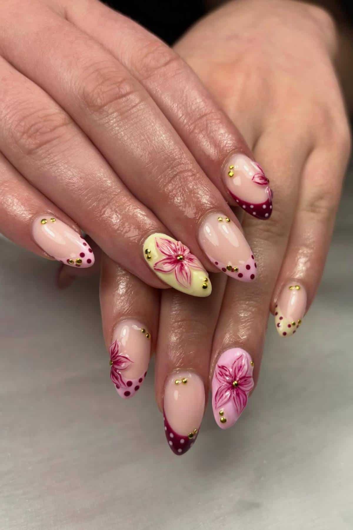 Hands with long almond-shaped nails featuring pink and yellow floral designs, gold studs, and burgundy polka-dotted tips on a light-colored surface.