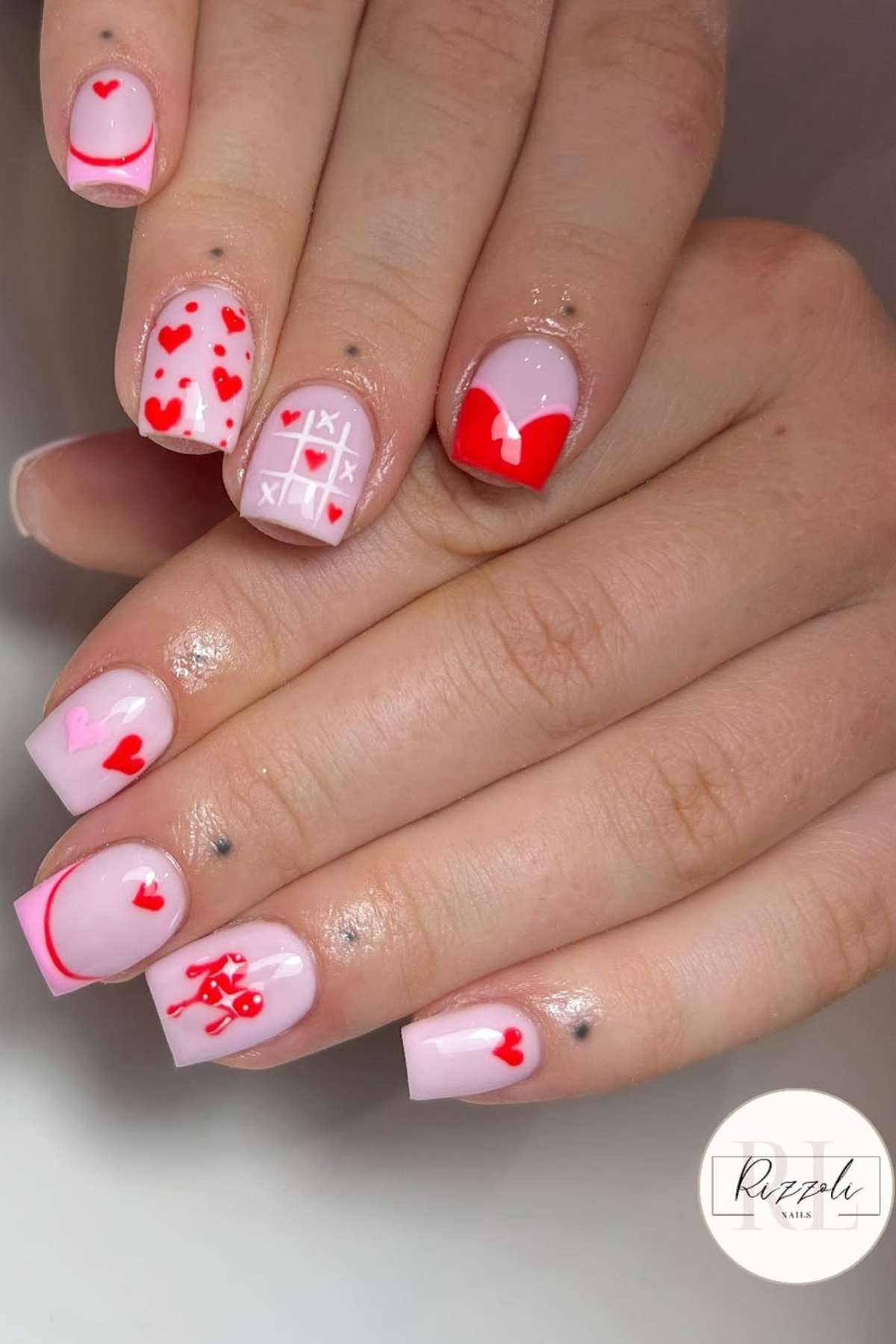 Hands with short, square-shaped nails painted light pink and decorated with red and pink heart, bow, tic-tac-toe, and line art designs.