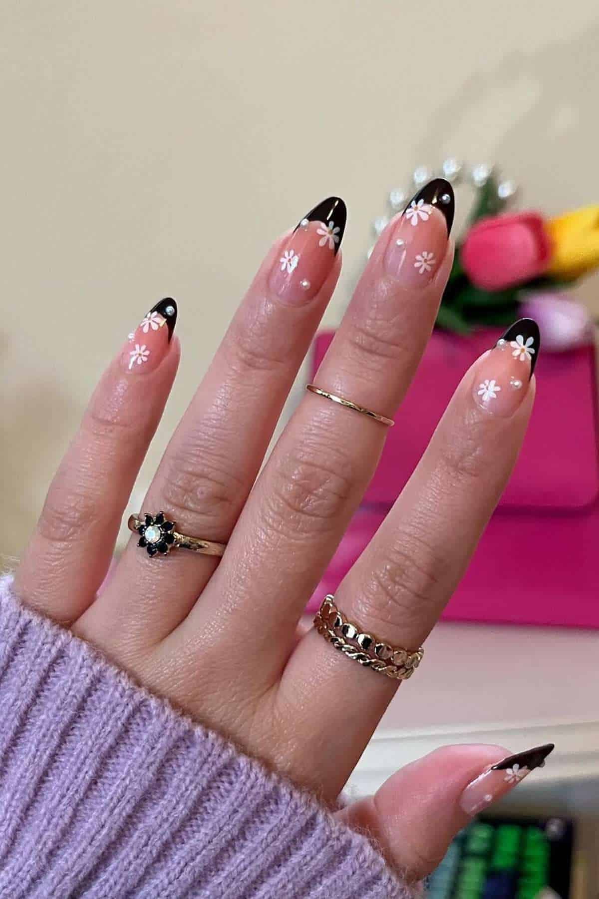 A hand with long, almond-shaped nails featuring black French tips and white daisy designs; the person wears three rings and a lavender sweater.