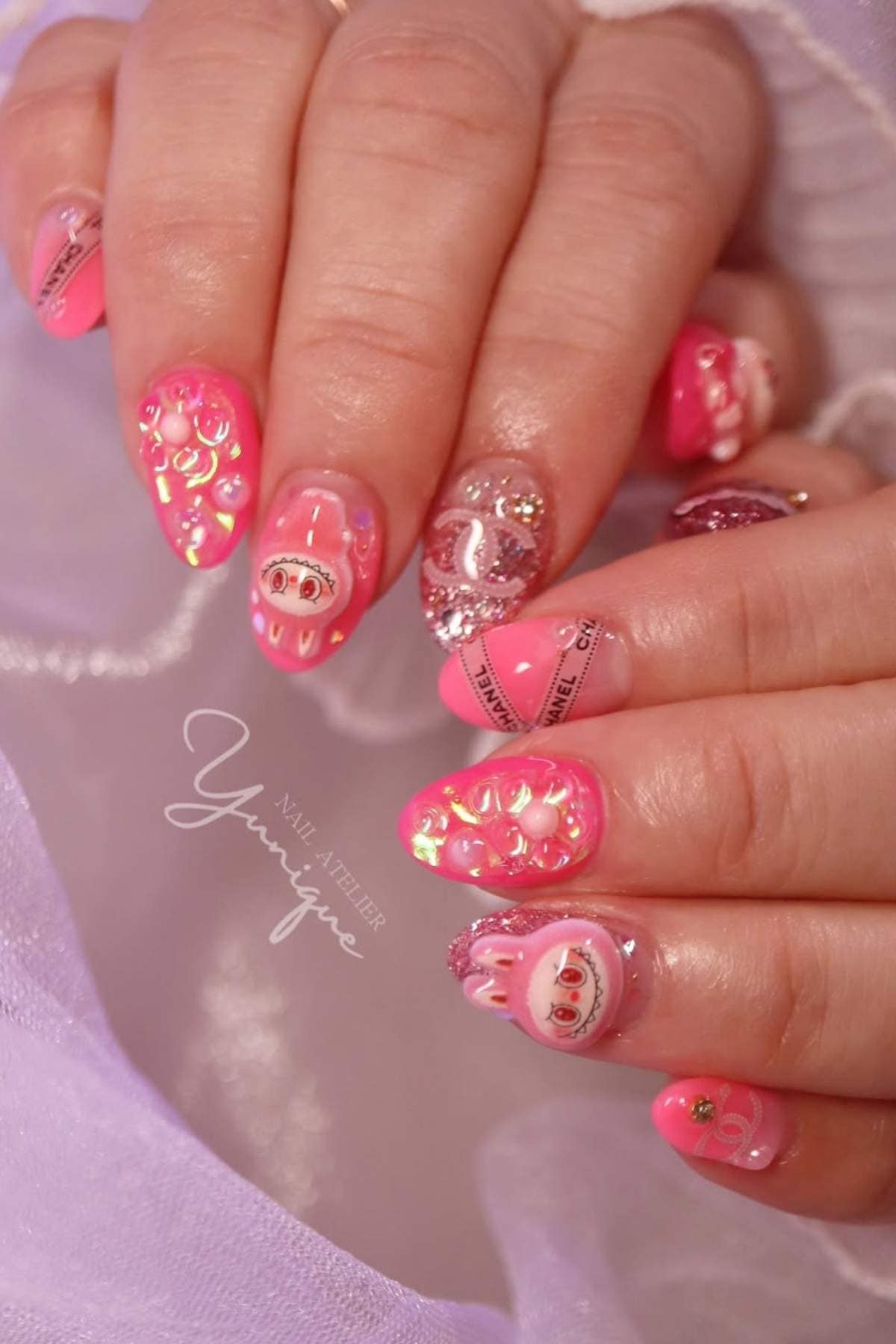 A hand with long, almond-shaped nails painted pink and red with 3D designs, glitter, and cartoon-like decorations.