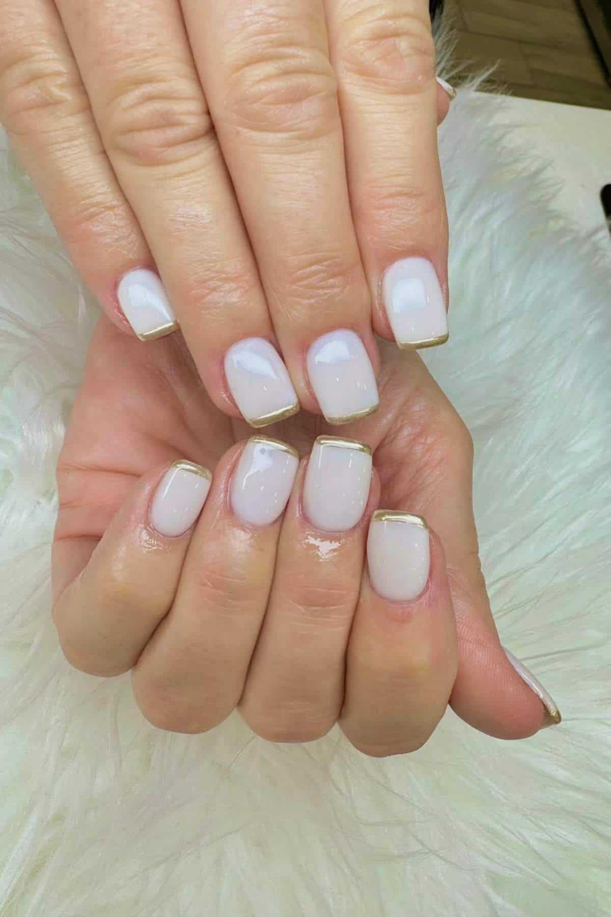 A close-up of hands with short, square-shaped nails painted light pink and thin gold tips, resting on a white, fluffy surface.