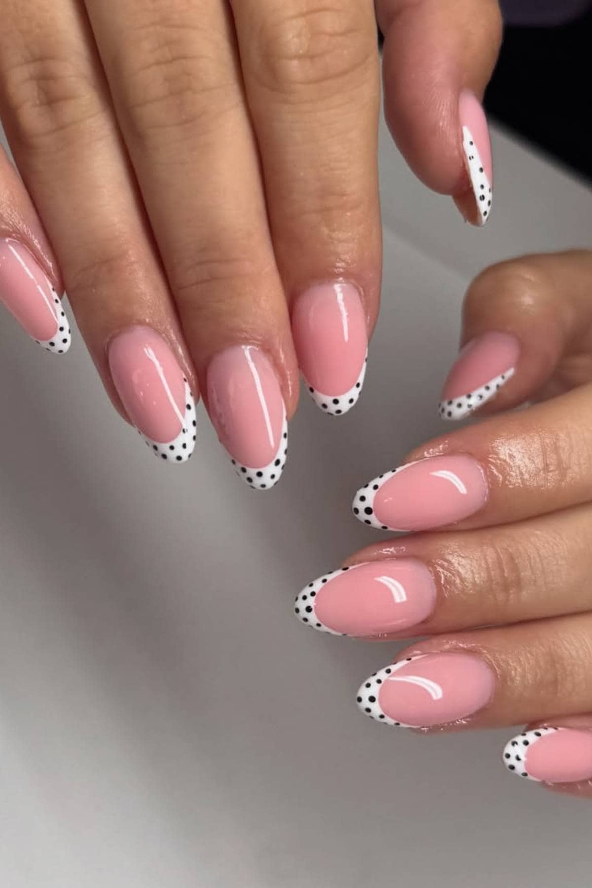 Hands with almond-shaped nails painted light pink, featuring white tips with black polka dots in a French manicure style.