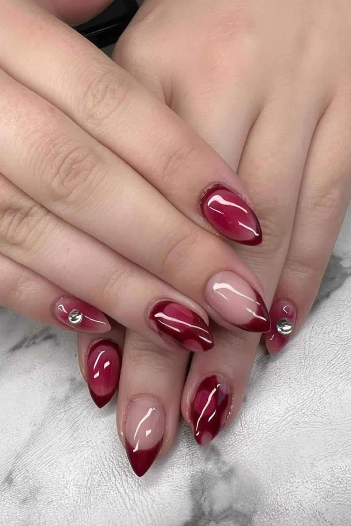 Hands with almond-shaped nails featuring a glossy red French tip manicure, some nails with rhinestone accents, on a marble surface.