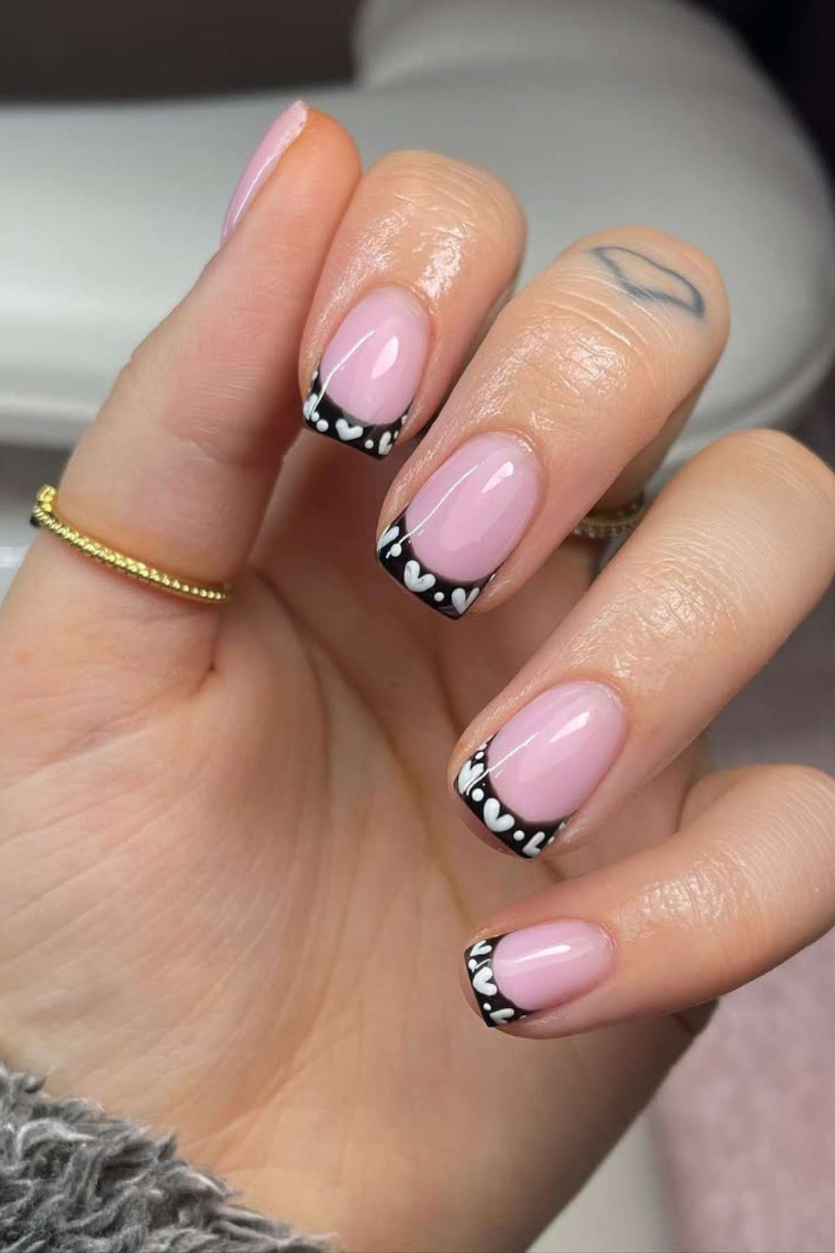 A hand with short, square-shaped nails painted light pink with black French tips and white heart designs, wearing gold rings and showing a lip tattoo on the finger.