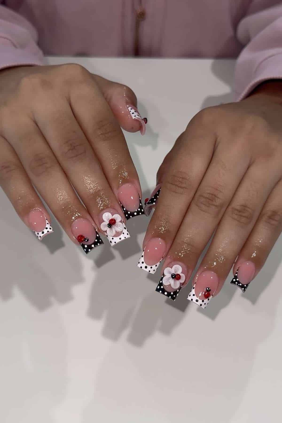Hands with long, square-shaped nails featuring pink bases, black and white polka dot French tips, 3D flower designs, and small ladybug decorations.