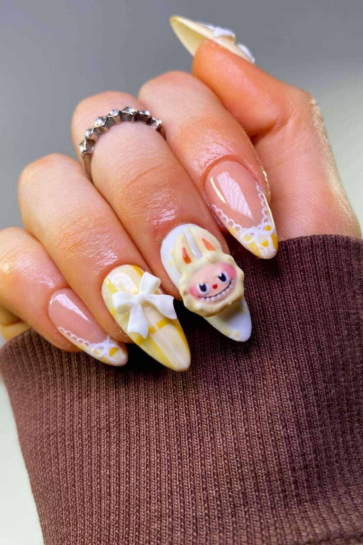 A hand with long, almond-shaped nails featuring yellow and white designs, a 3D bow, and a 3D bunny face accent nail. The person wears a silver ring and a brown sleeve.
