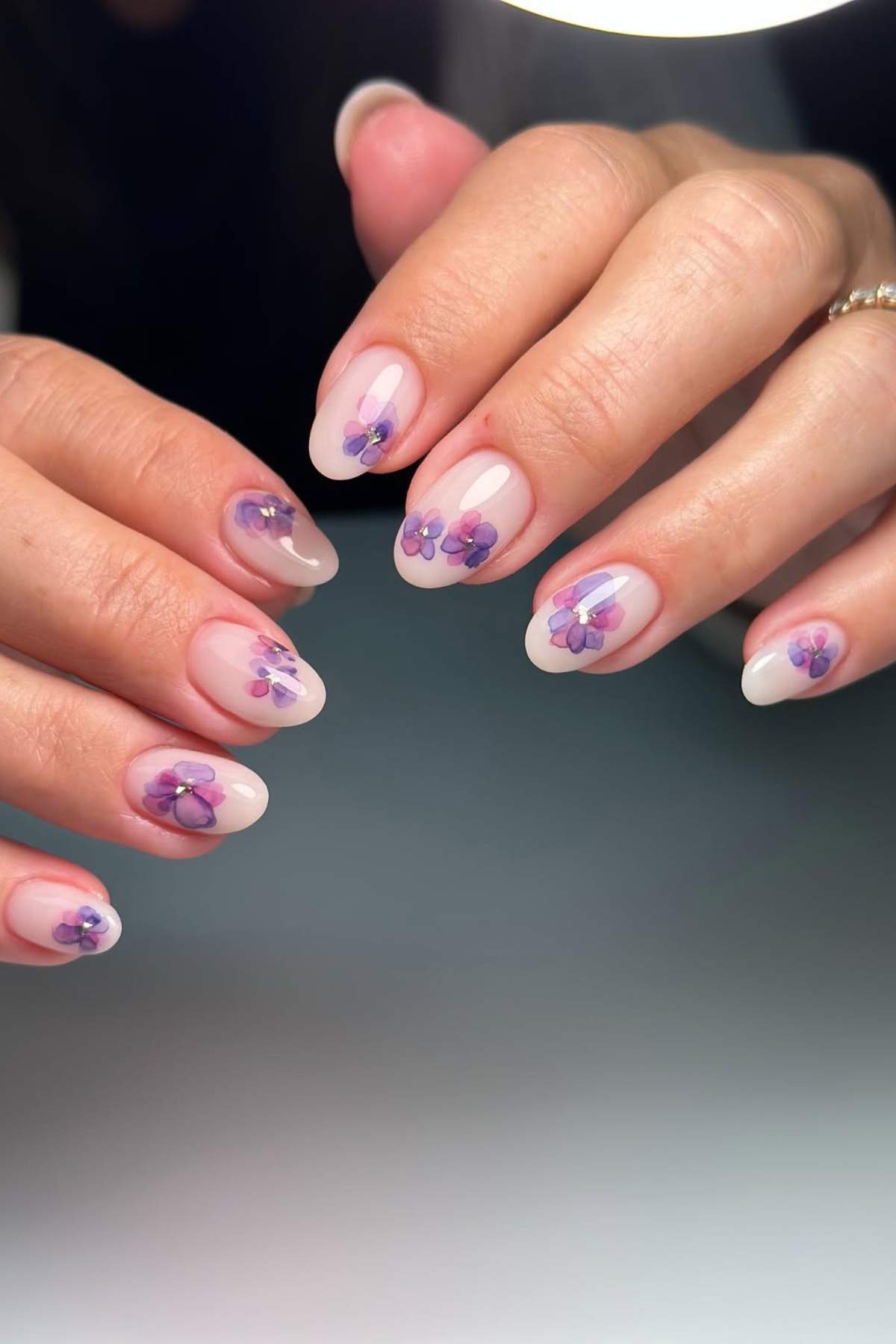 Hands with almond-shaped nails featuring a light pink base and purple floral nail art design on each nail.