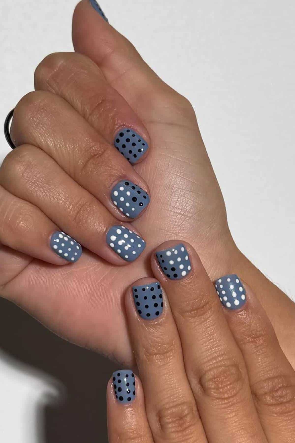 Hands with short, matte blue nails featuring alternating patterns of white and black polka dots on each nail.