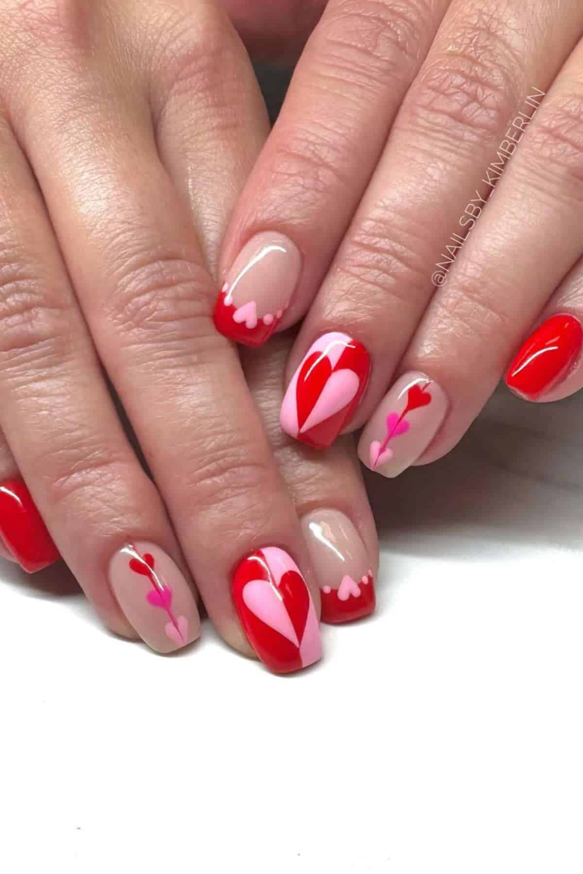 Hands with manicured nails featuring red and pink heart-themed nail art designs on a nude and red base.