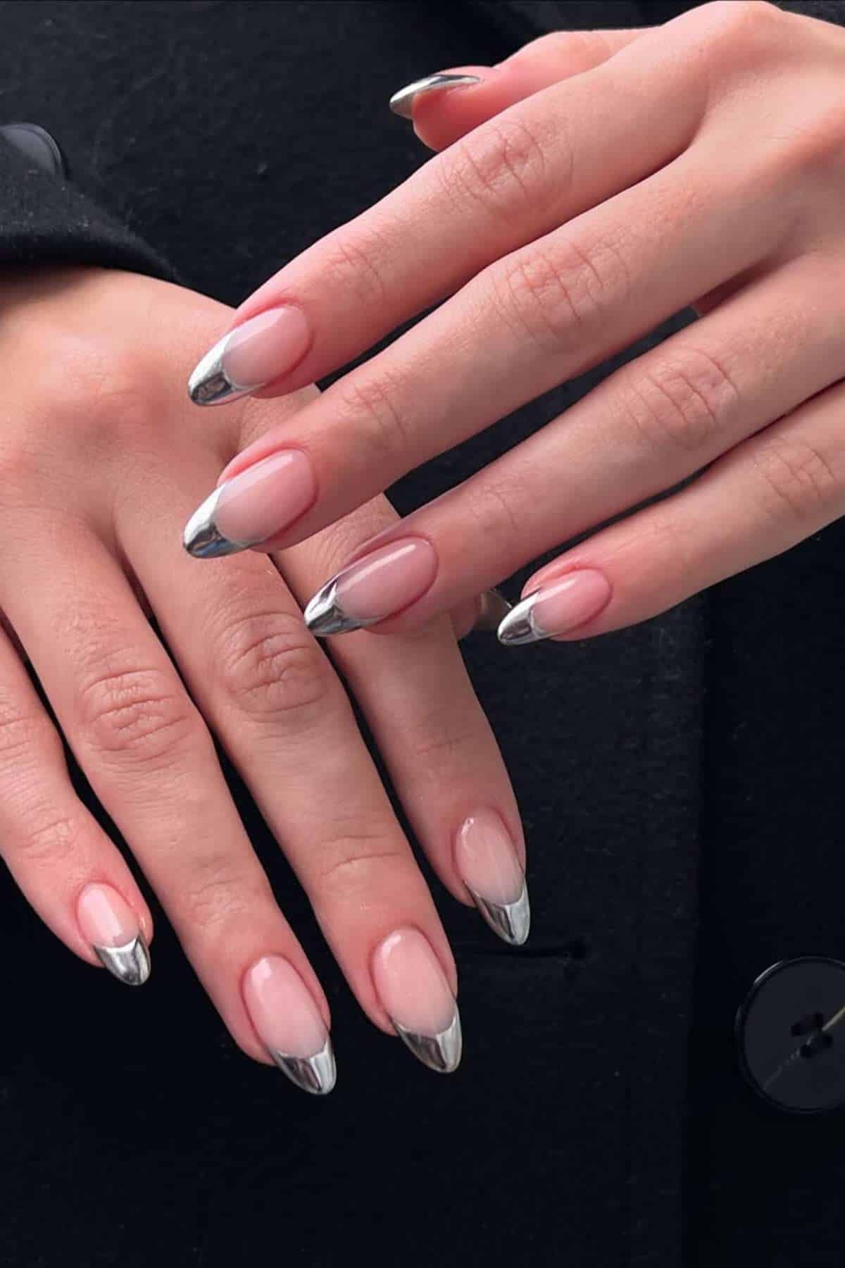 Close-up of hands with long almond-shaped nails featuring a nude base and metallic silver tips, resting on a black coat.