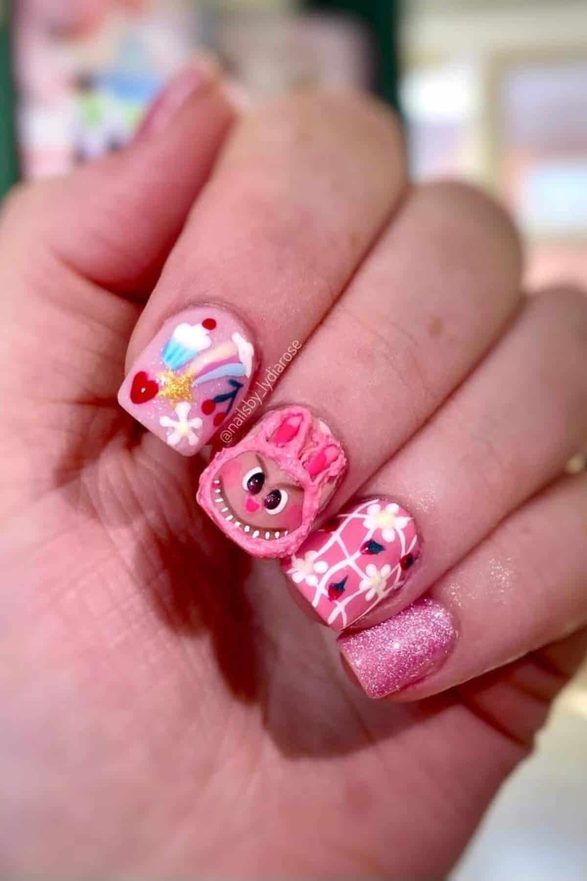 A hand with pink-themed nail art featuring flowers, hearts, and a three-dimensional bear face design on the middle finger.
