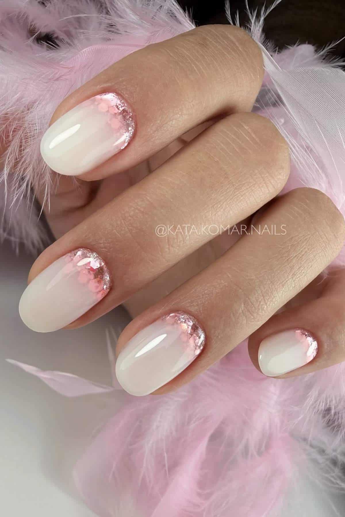 A hand with short, oval-shaped nails featuring a pale pink base and pink glitter accents near the cuticles, surrounded by soft pink feathers.
