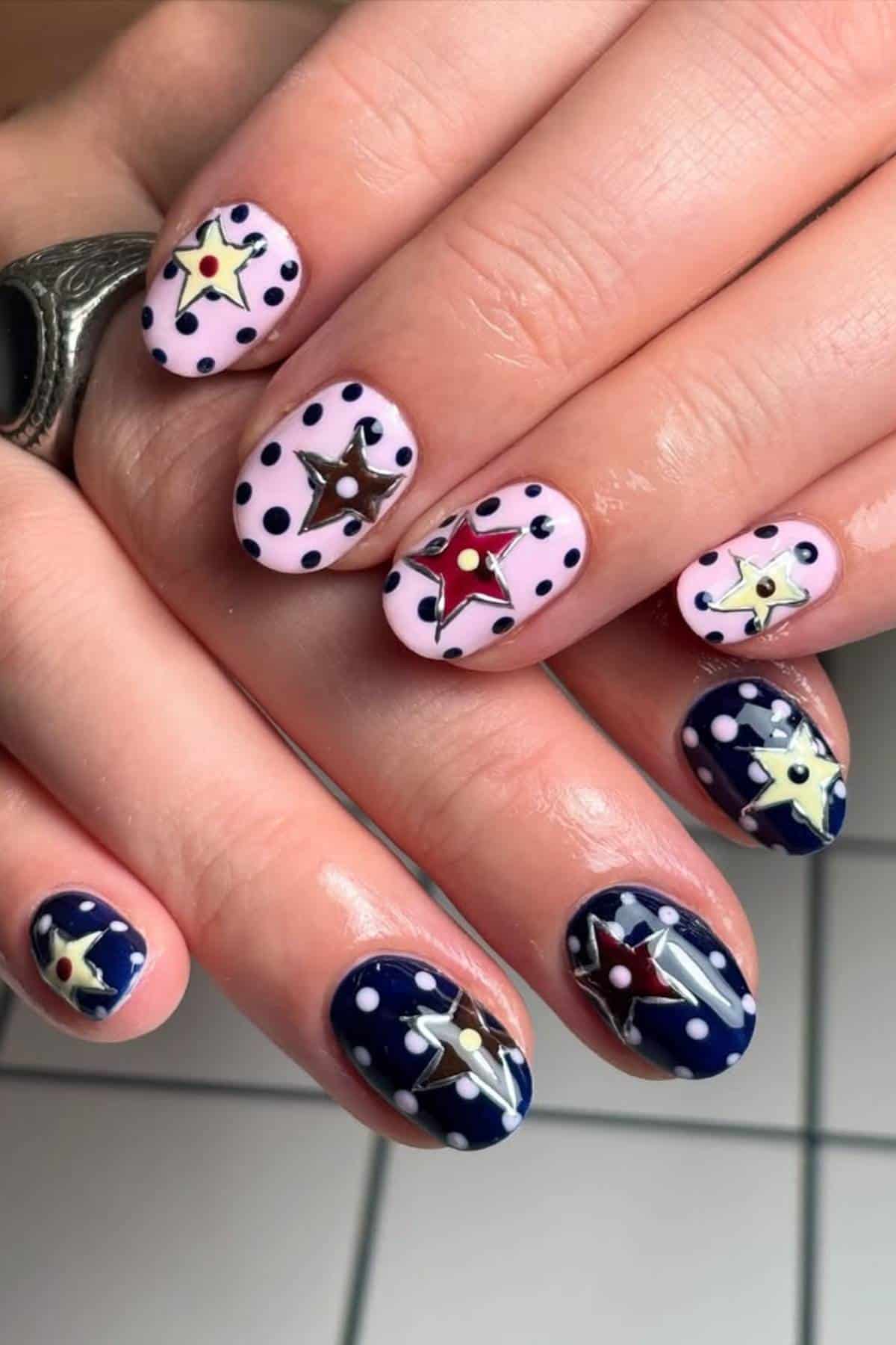 Hands with manicured nails featuring pink and navy blue bases, decorated with polka dots and star-shaped designs in red, yellow, and gold.
