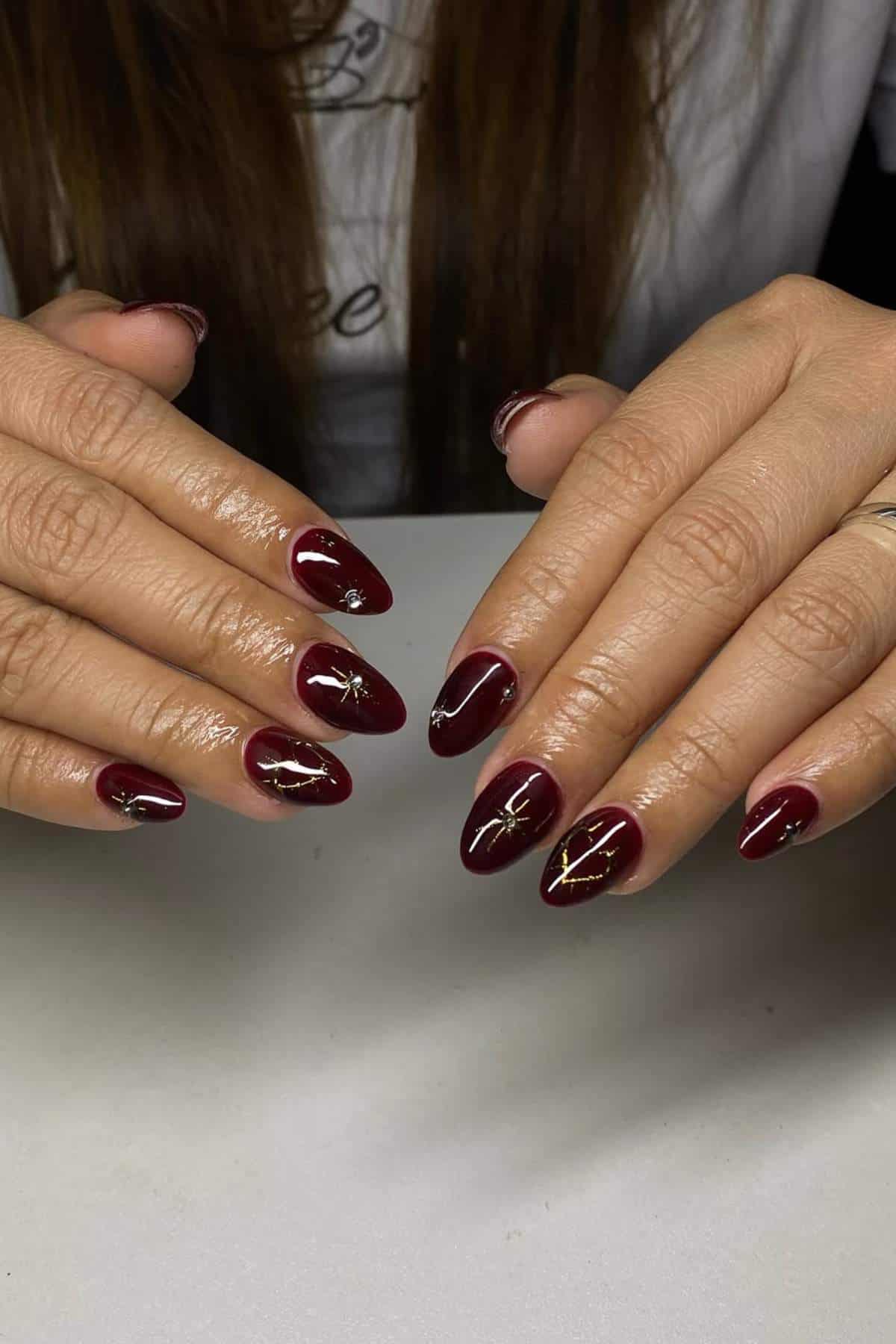 Hands with long, almond-shaped nails painted dark red with gold foil accents and rhinestone details, resting on a white surface.
