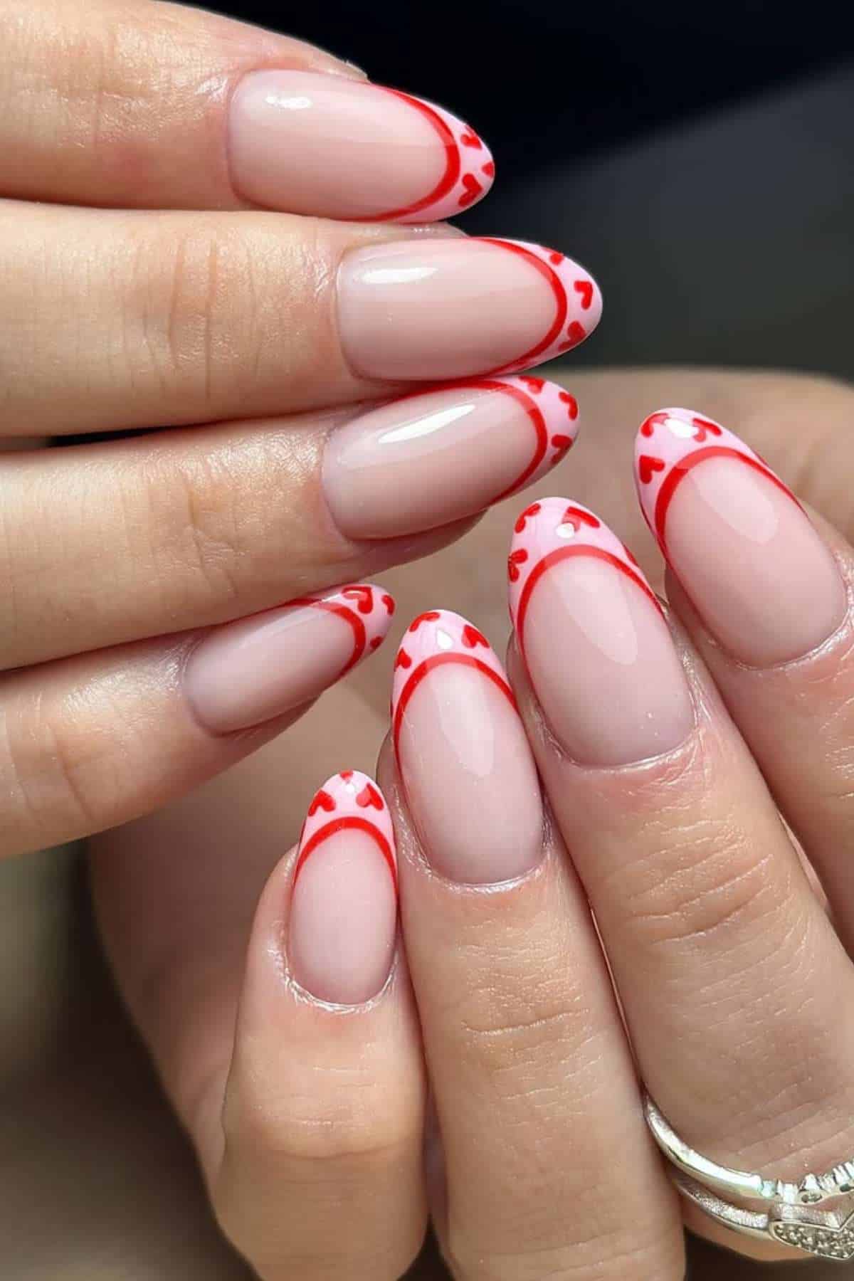 Close-up of hands with almond-shaped nails featuring a nude base and red heart-shaped French tip designs.