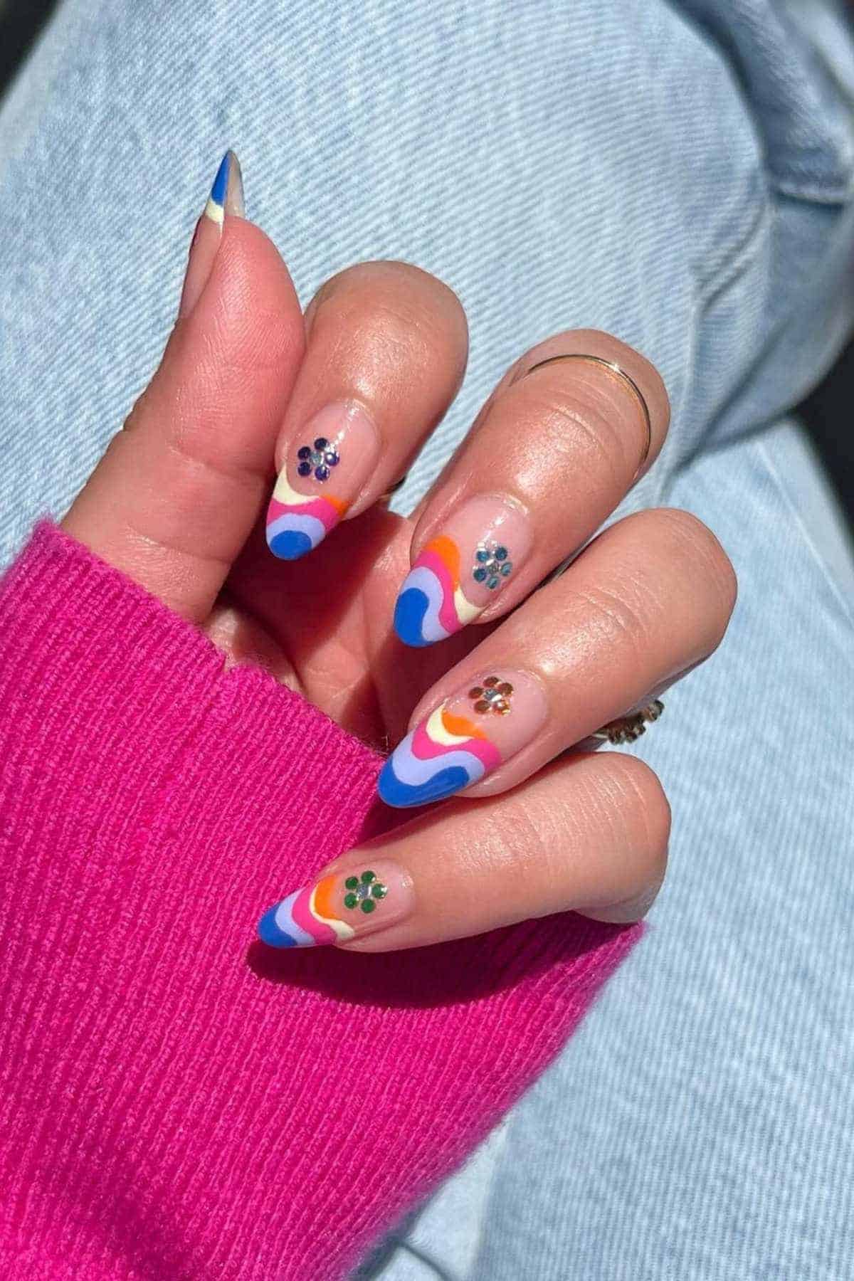 A hand with almond-shaped nails featuring colorful wavy patterns and small flower decals, wearing a pink sleeve and blue jeans.