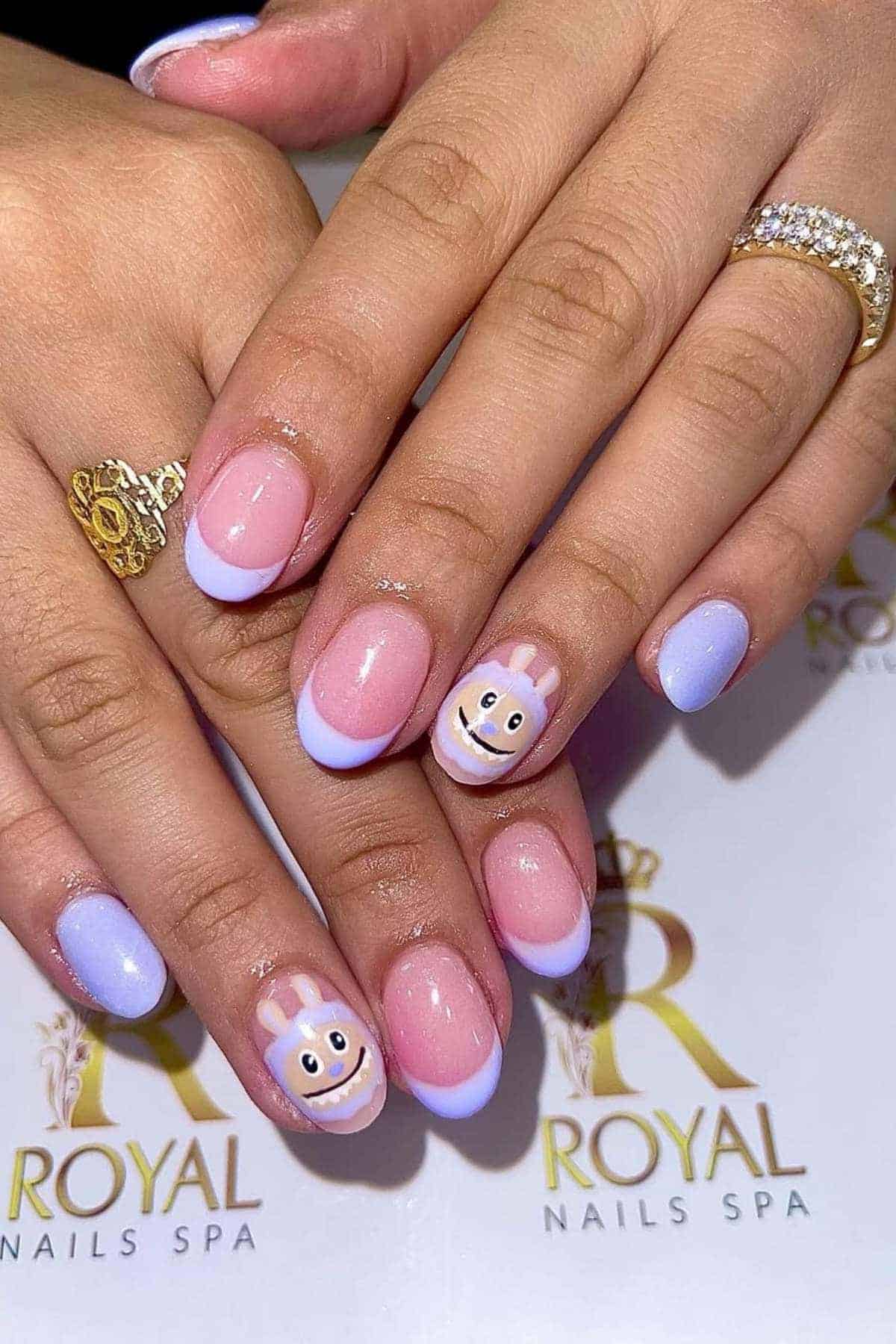 Hands with pastel purple French tip manicure; two nails feature smiley face designs with bunny ears. The person is wearing a gold ring with clear stones.