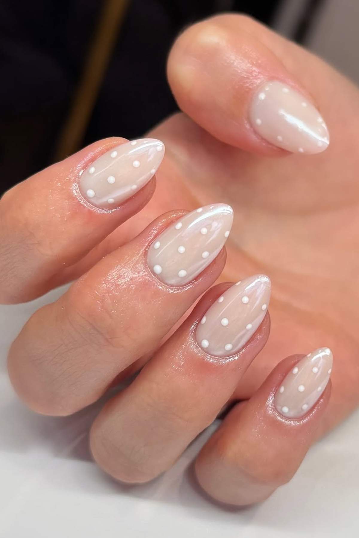 A hand with almond-shaped nails painted a sheer nude color and decorated with white polka dots.