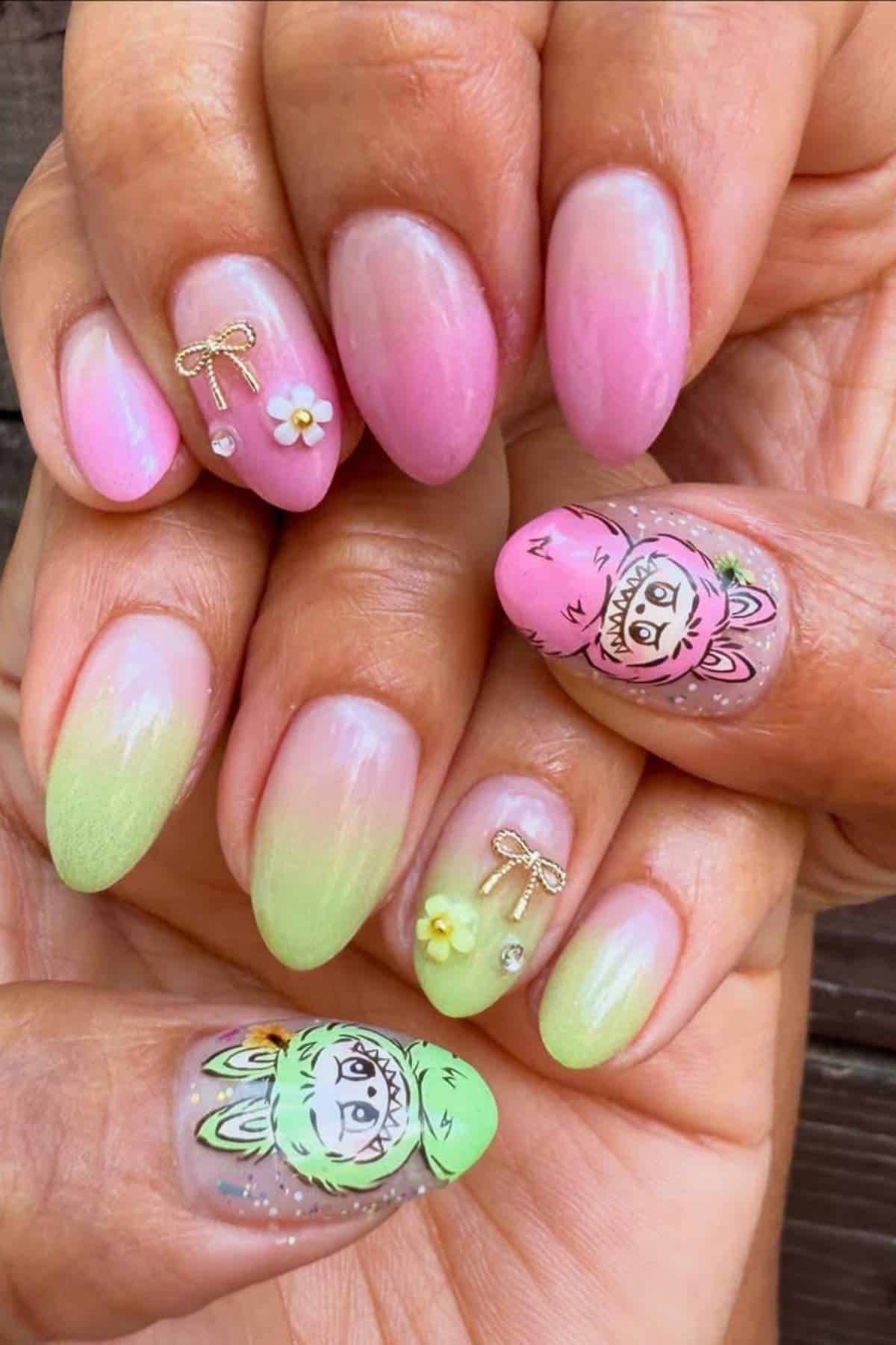 Hands with almond-shaped nails featuring pink and green ombre polish, gold bows, small flower charms, rhinestones, and cartoon animal face nail art on the thumbs.