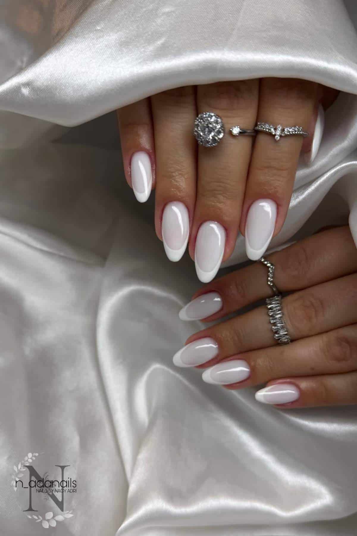Hands with almond-shaped French manicure in white, wearing silver rings, resting on white satin fabric.