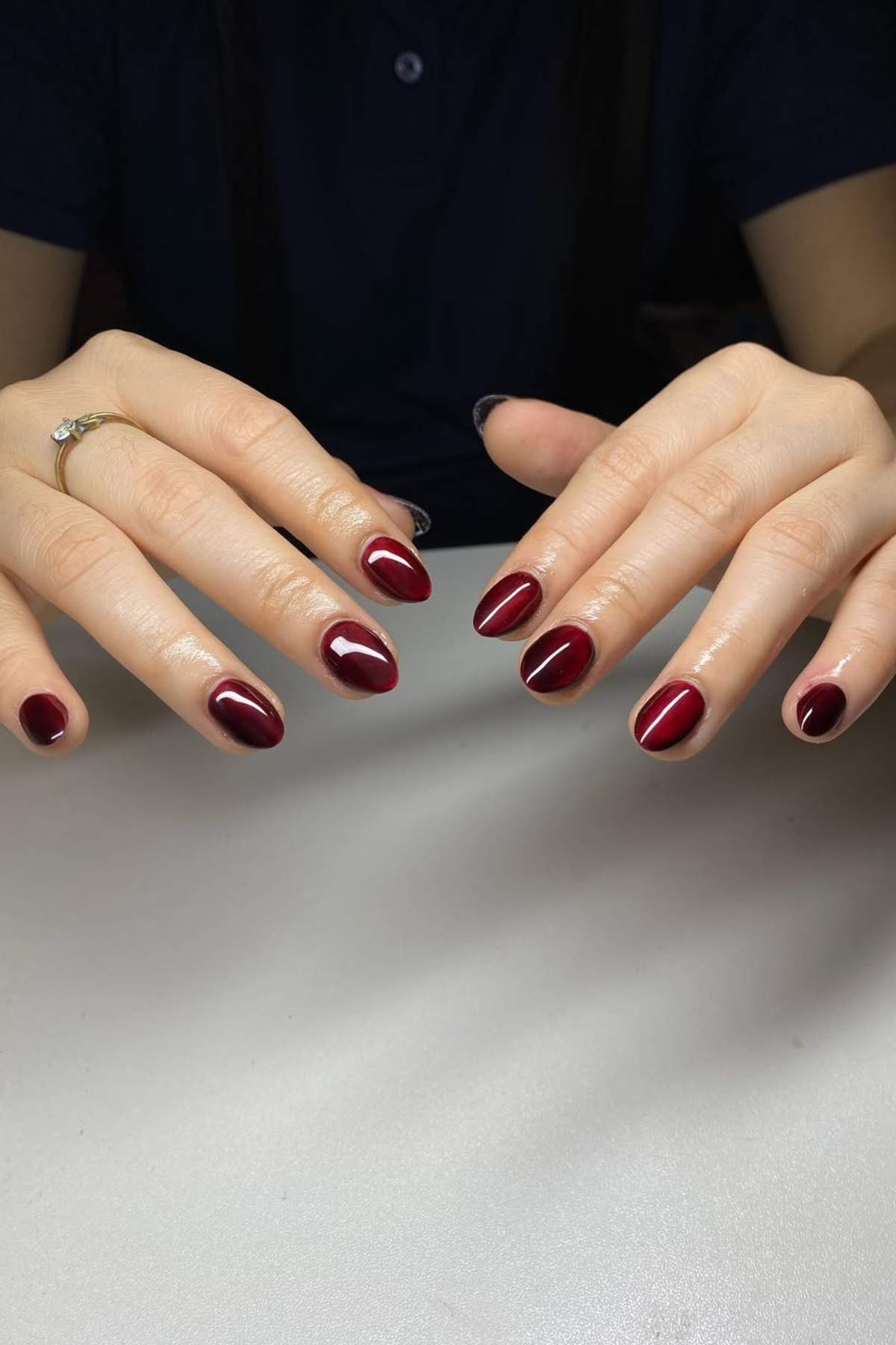 Two hands with almond-shaped fingernails painted in glossy dark burgundy nail polish, positioned over a light surface.
