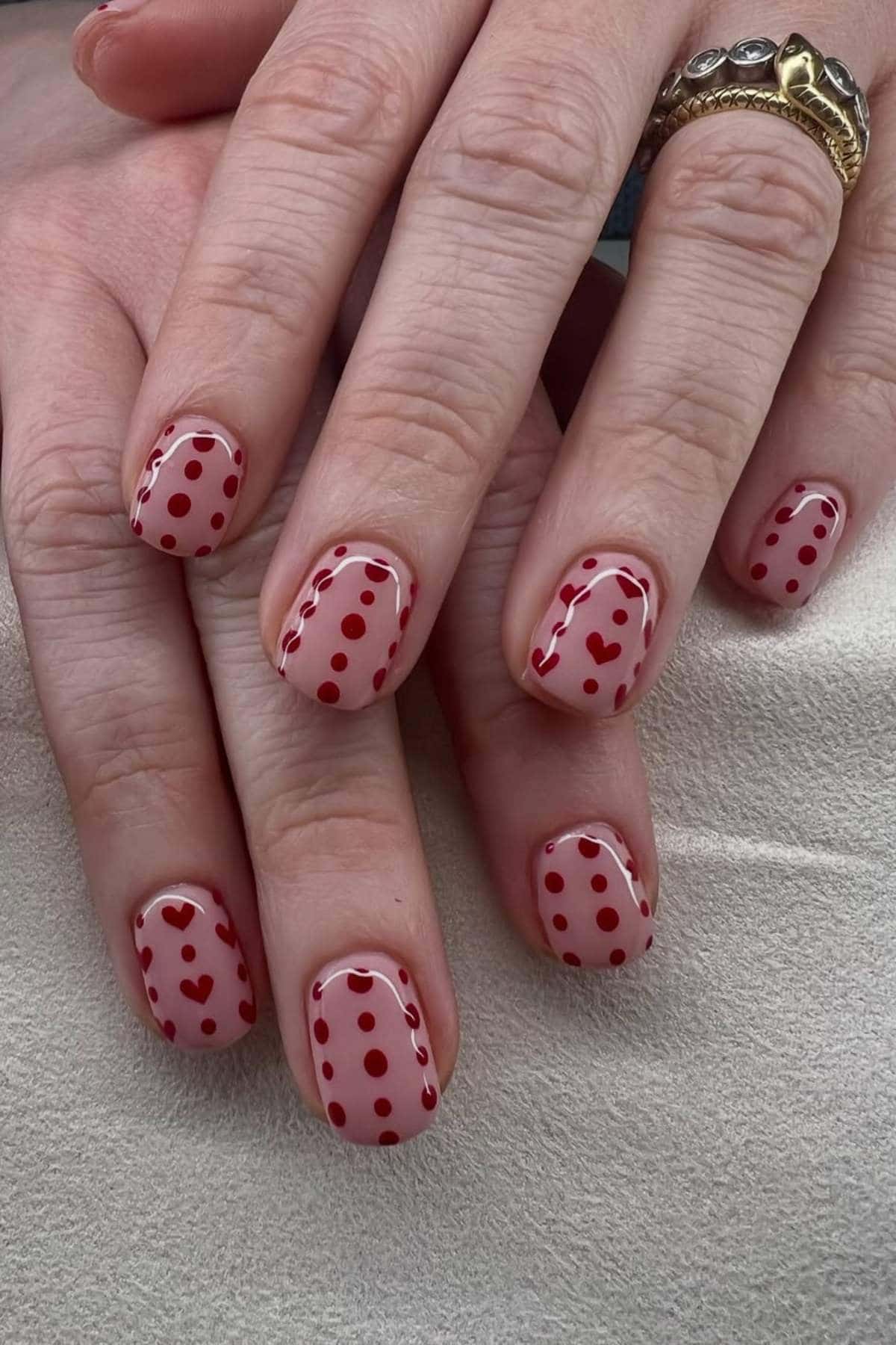 Hands with short, nude-colored nails featuring red polka dots, hearts, and wavy lines as nail art. The person is wearing gold rings.