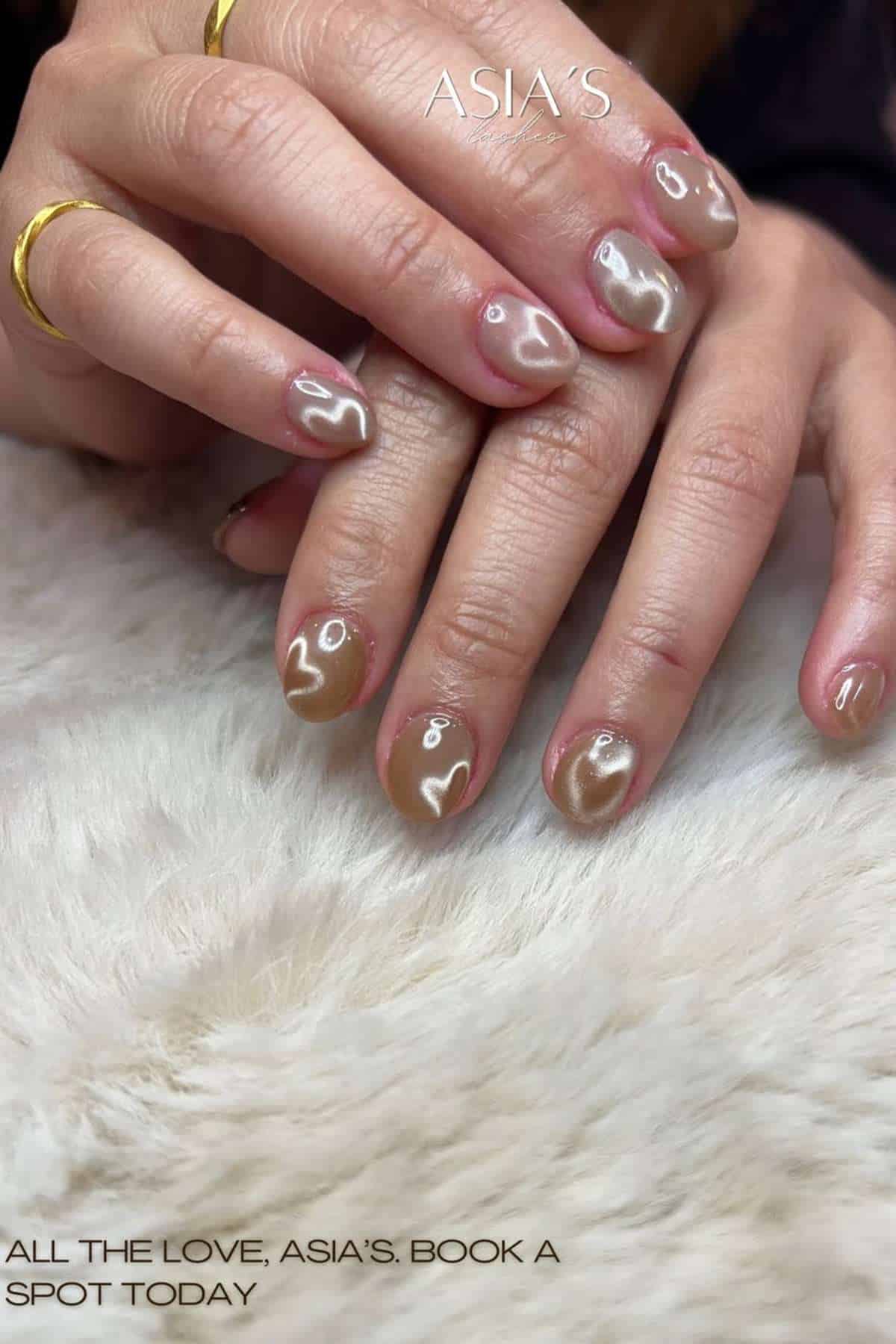 Close-up of hands with brown and beige heart-shaped nail art, wearing gold rings, resting on a white textured surface. Text reads: "ALL THE LOVE, ASIA'S. BOOK A SPOT TODAY.