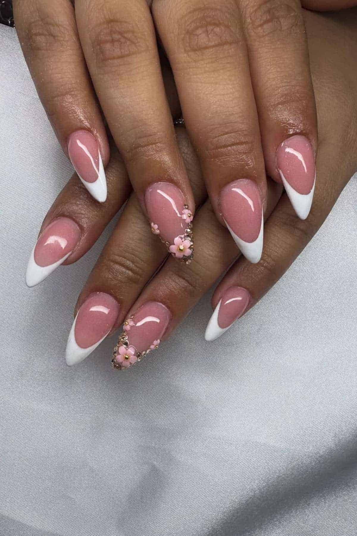 Hands with almond-shaped nails featuring a pink base, white French tips, and 3D pink floral decorations on two accent nails, displayed on a white surface.