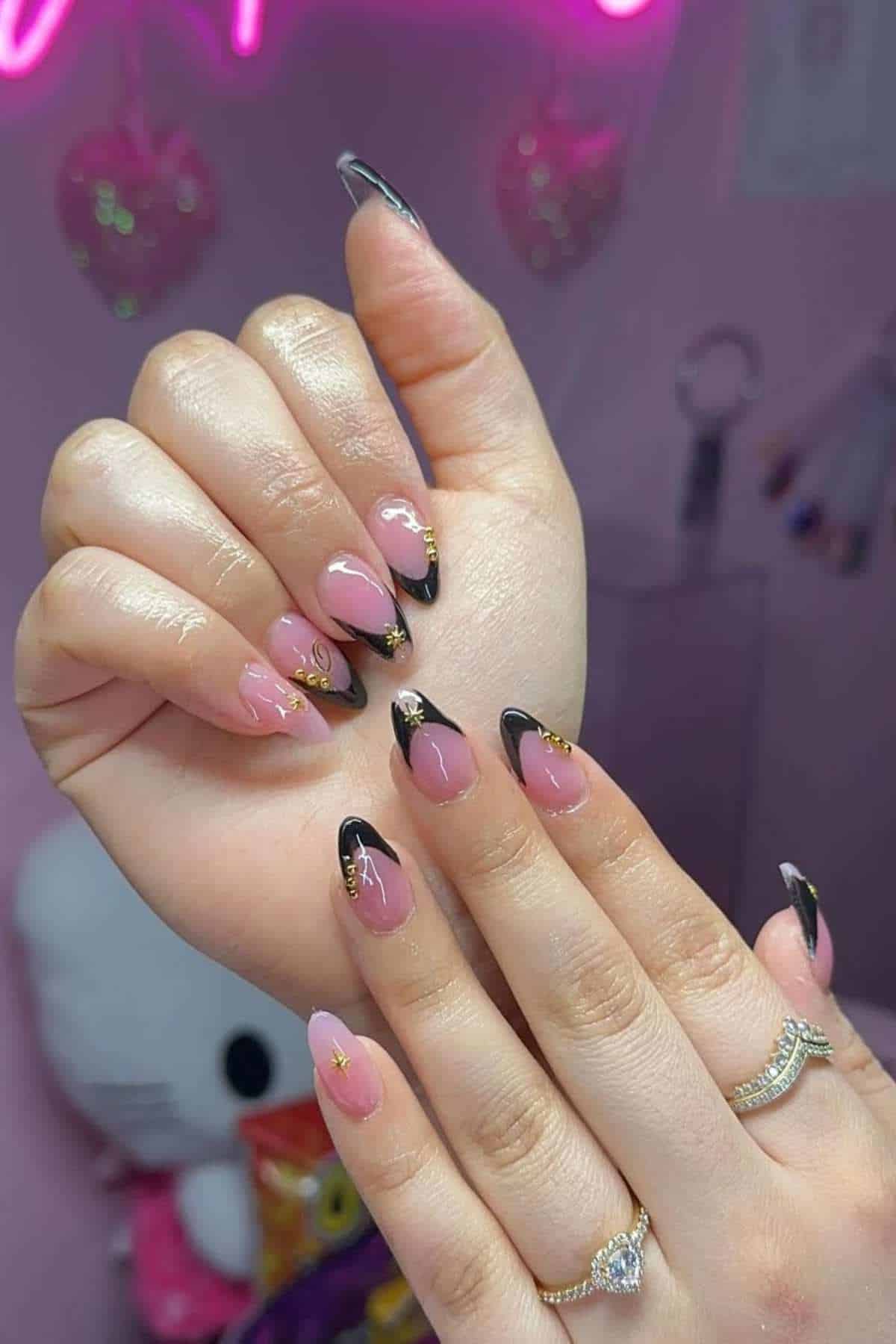 A person displays manicured nails with a pink base, black tips, and gold accents, wearing rings on both hands against a pink background.