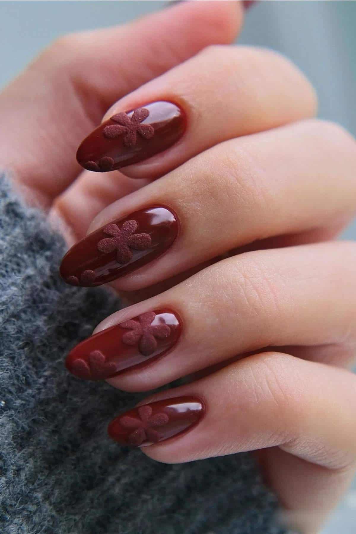 A hand with almond-shaped nails painted glossy burgundy, featuring raised matte floral designs on each nail. The person is wearing a gray knit sweater.