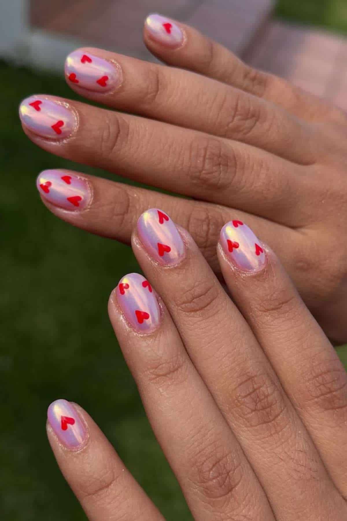 Two hands with short, oval nails painted in a light, iridescent base color and decorated with small red heart designs on each nail.