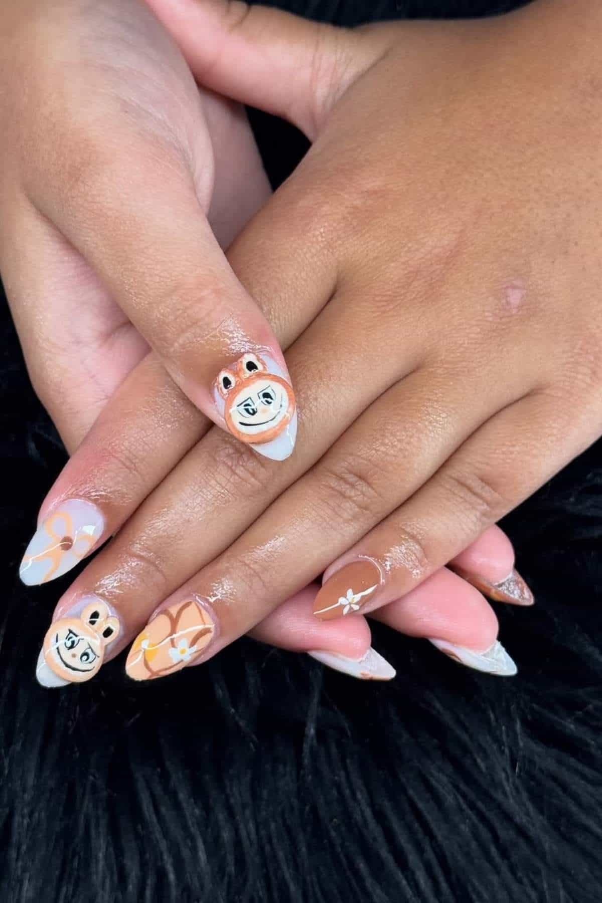 Hands with long, almond-shaped nails featuring cartoon bear faces, floral designs, and brown, white, and beige color patterns, resting on a black furry surface.