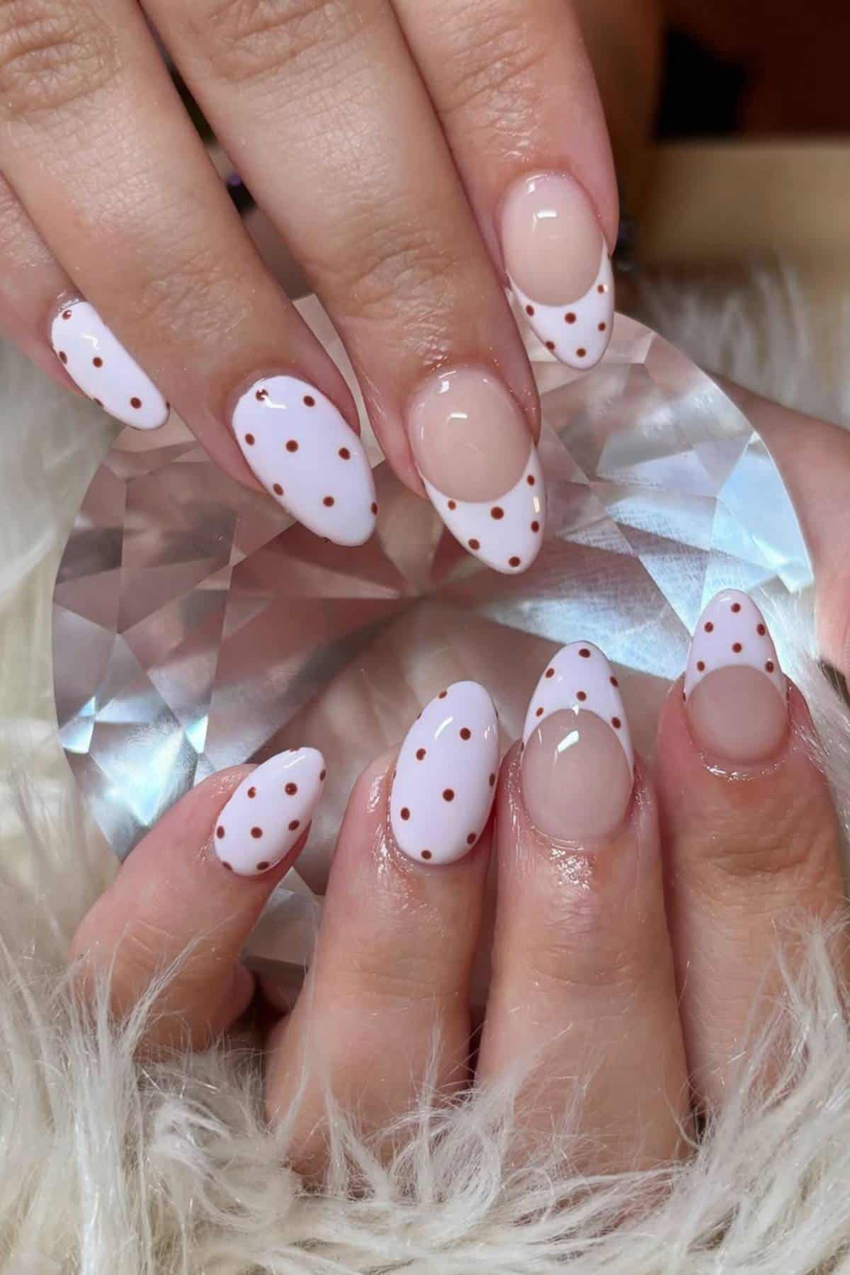 Hands with almond-shaped nails featuring a white French tip design and small brown polka dots, holding a faceted crystal object, with a furry white surface underneath.