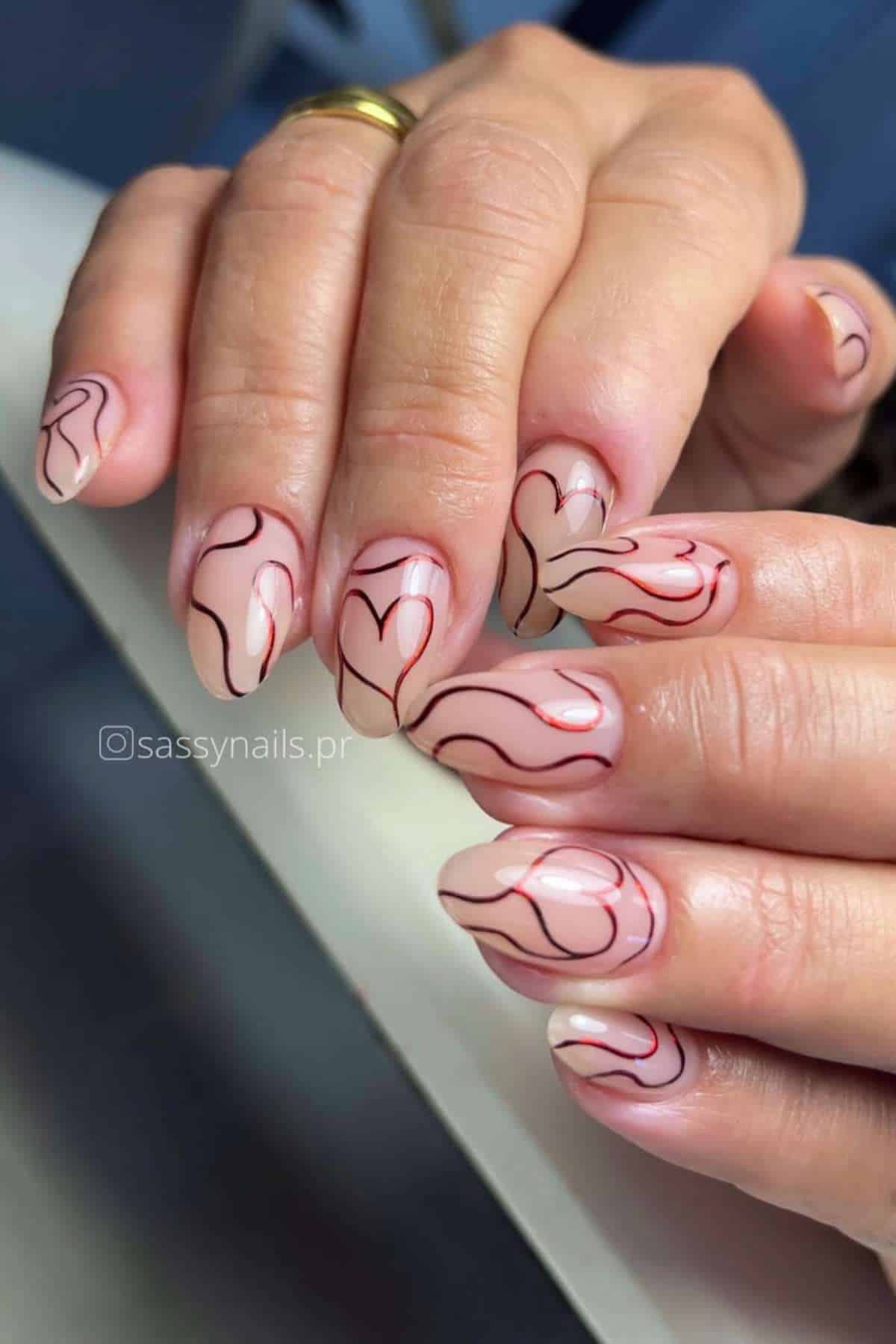 Close-up of hands with nude-colored nails featuring abstract red and black line art designs, including heart shapes.