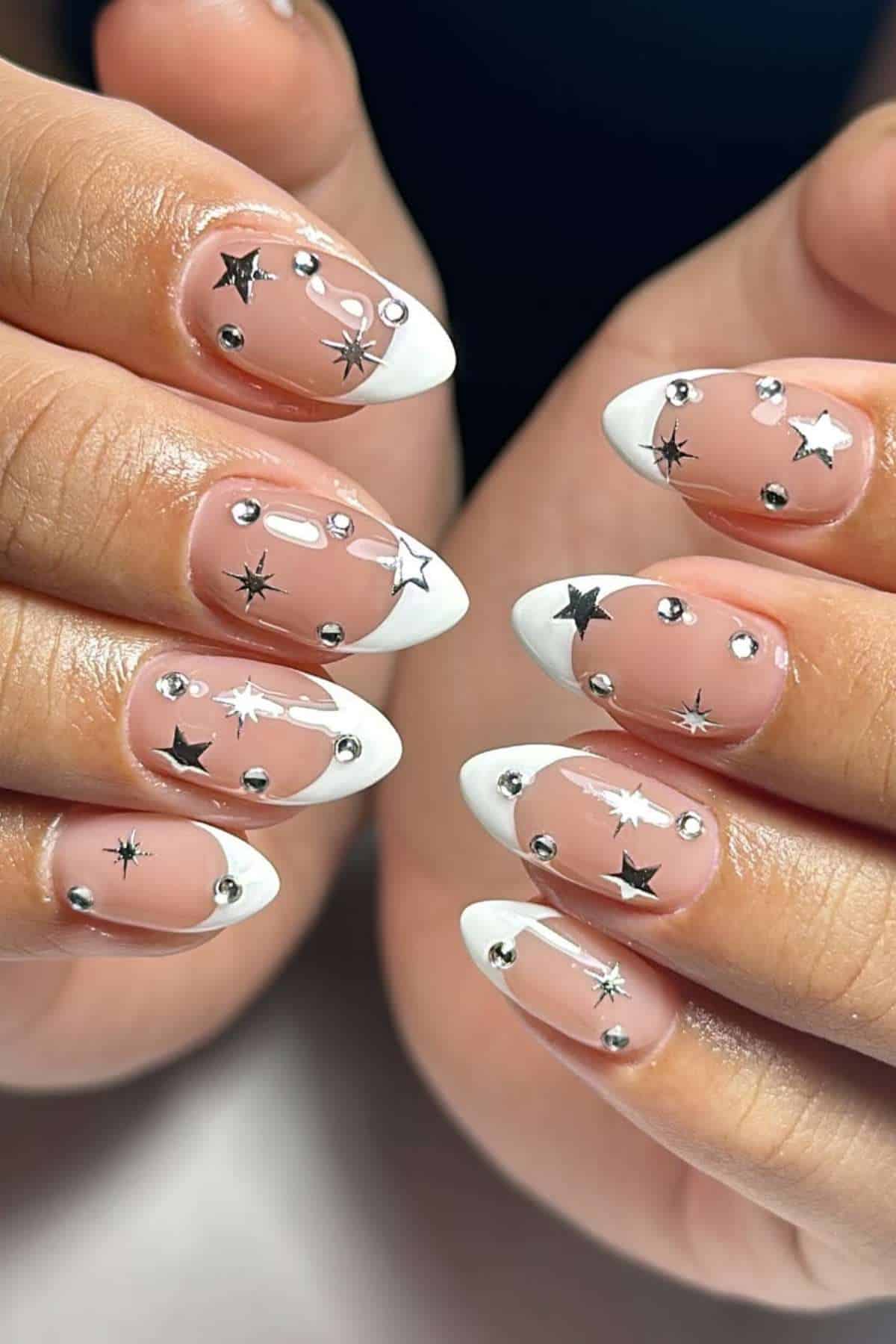 Hands with almond-shaped nails featuring white French tips, silver studs, and black star designs on a nude pink base.