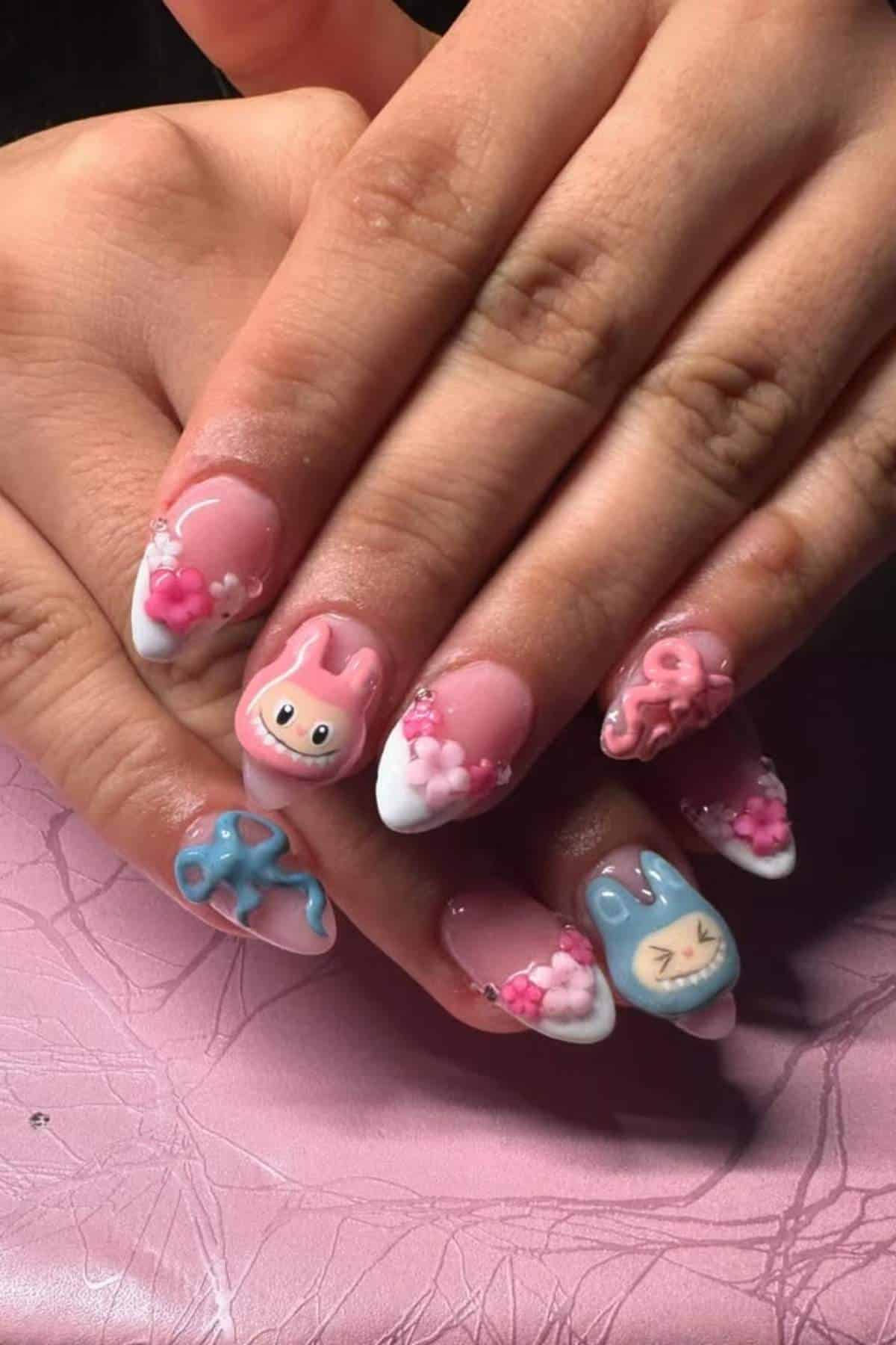 Hands with long, almond-shaped nails featuring 3D pastel bunny and octopus designs, pink and blue colors, and small flower decorations on a pink textured surface.