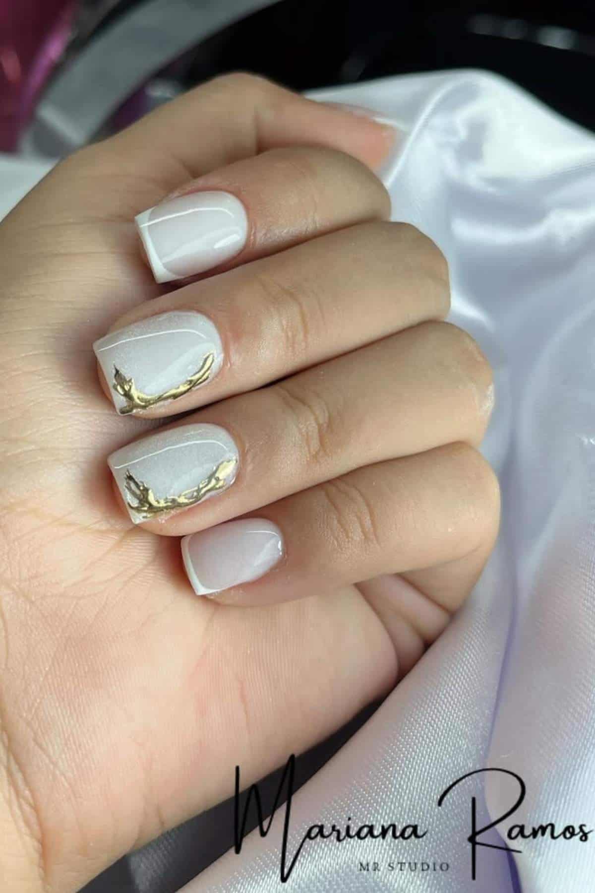 A hand with short, square-shaped white nails featuring a gold swirl design on the ring and middle fingers, resting on white satin fabric.