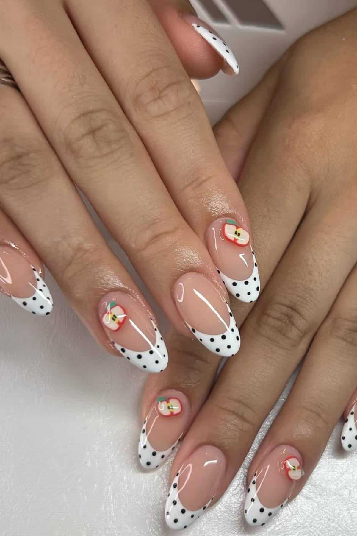 Hands with almond-shaped nails featuring white tips with black polka dots and small red and yellow floral designs on some nails.