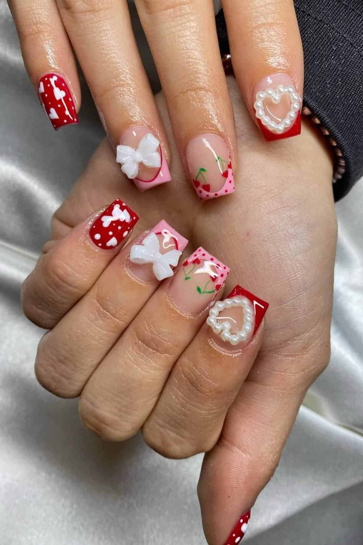 A hand with square-shaped nails featuring red and pink designs, white bows, hearts, polka dots, and floral accents.