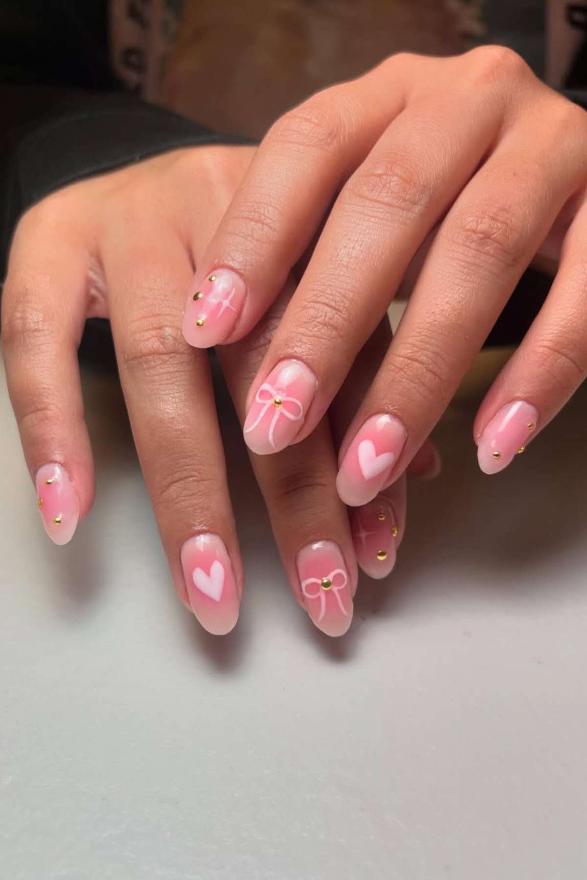 Close-up of hands with manicured nails featuring pink polish, white heart and bow designs, and small gold studs on a light surface.