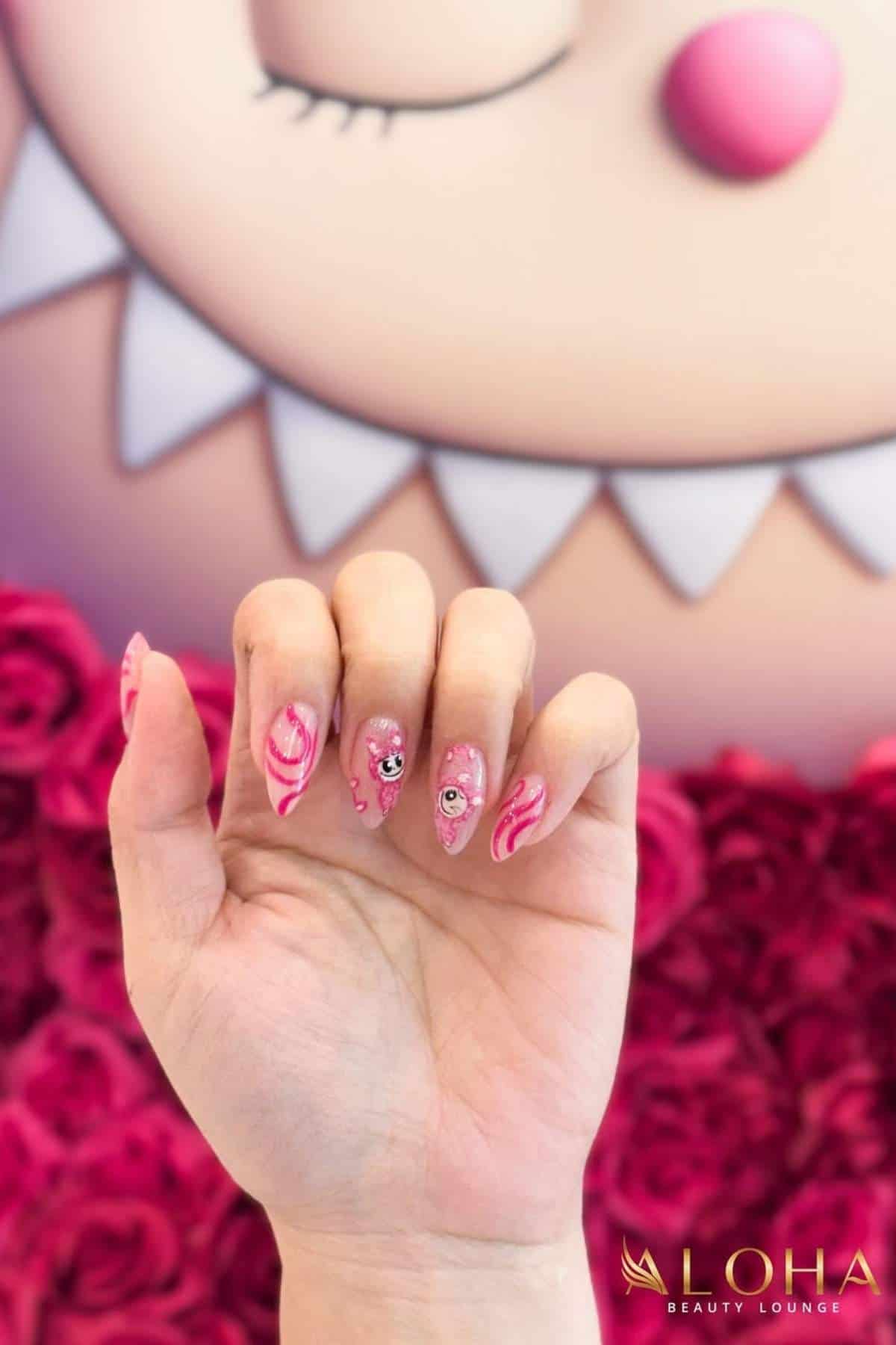 A hand with long pink and white marble nail art and small embellishments is held up in front of a pink cartoon face and roses. Logo for LOHA Beauty Lounge is visible in the corner.