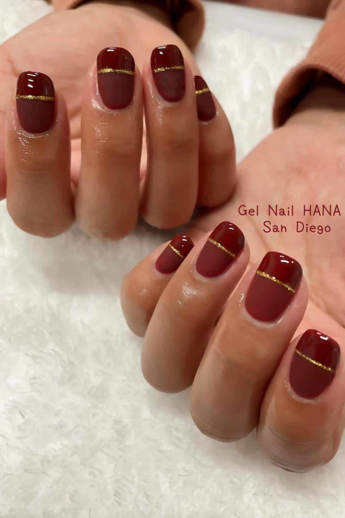 Hands with short, oval-shaped burgundy gel nails featuring a horizontal gold glitter stripe near the tips, displayed against a white background.