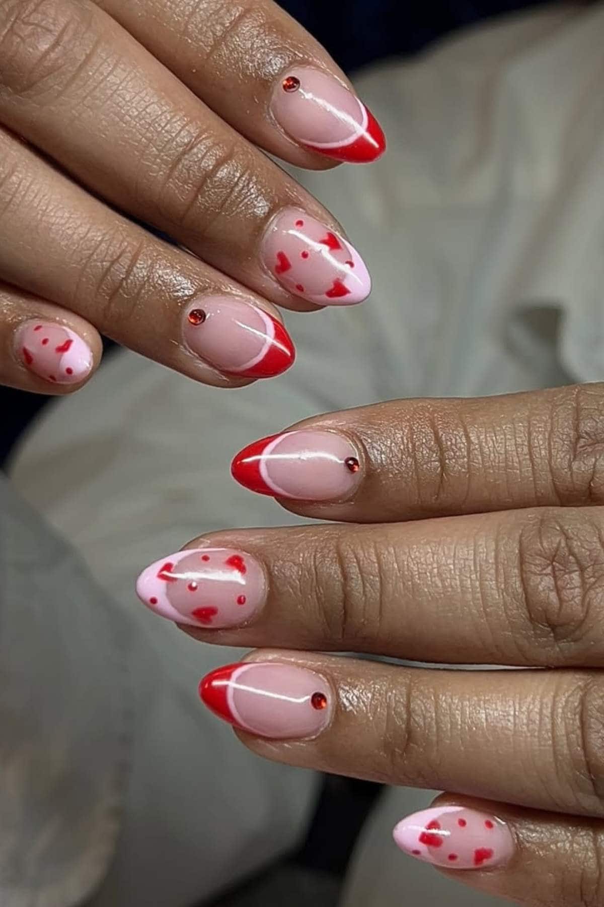 A close-up of hands with almond-shaped nails, featuring pink and red heart designs, red French tips, and small red rhinestone accents on each nail.